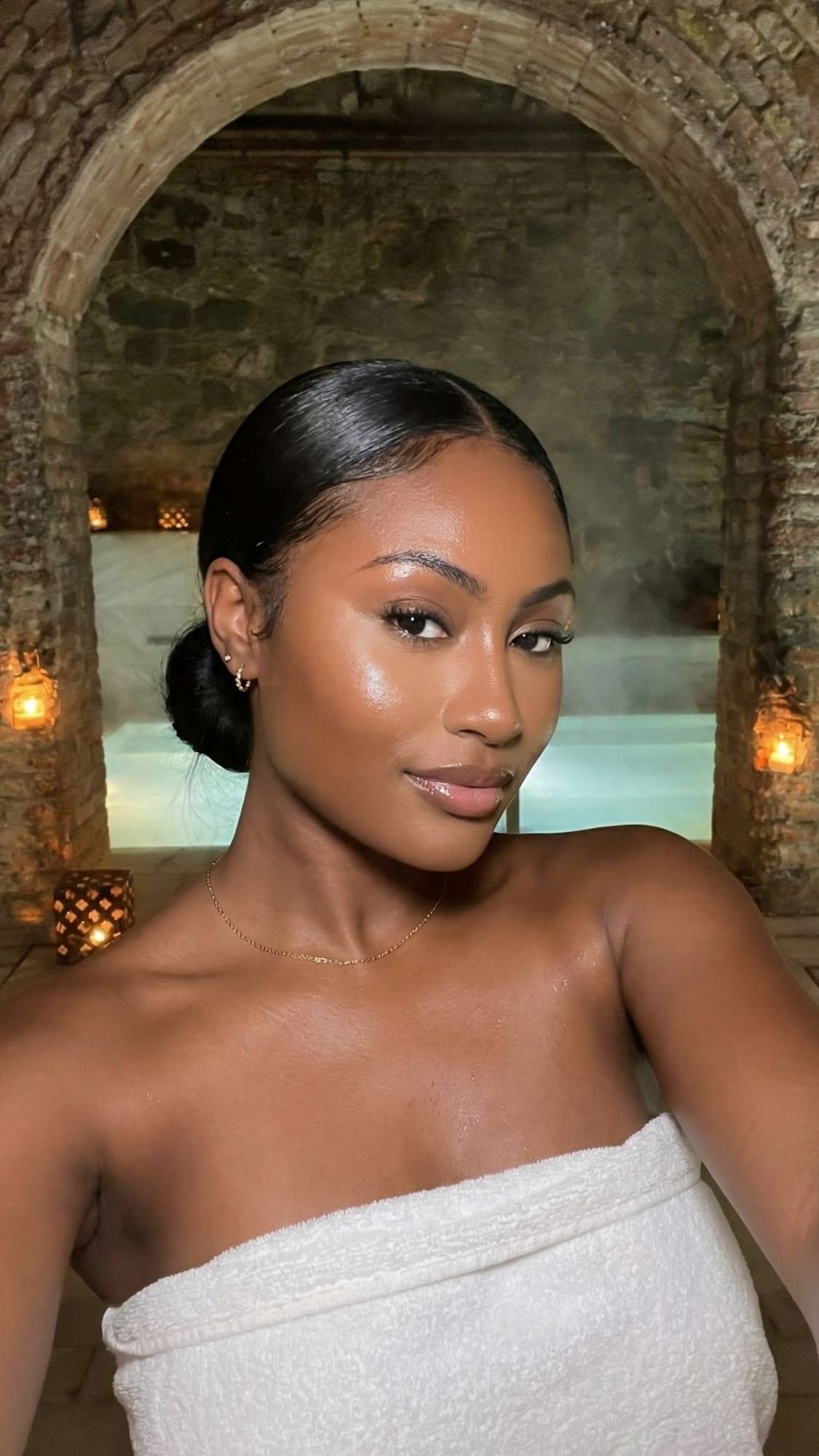 A woman with smooth, glowing skin and dark hair in a low bun takes a selfie in front of a stone archway and softly lit indoor pool—perfect for cozy winter plans during the off season. She wears a white towel and subtle makeup, with warm lantern lights behind her.