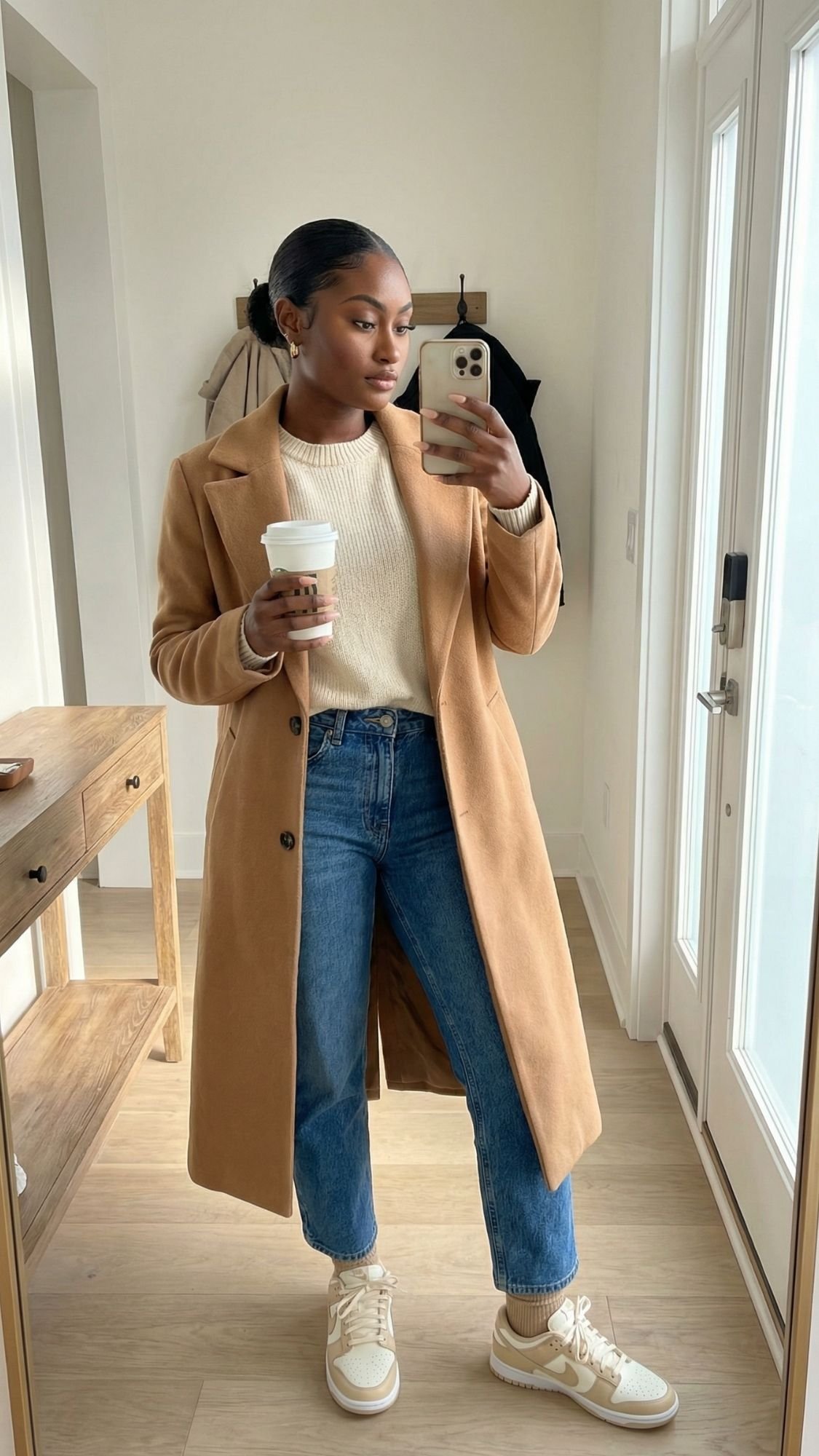 A woman stands indoors taking a mirror selfie, dressed in a camel-colored coat over a cream sweater, blue jeans, and white sneakers. Perfect for calm mornings, her outfit captures the essence of quiet girl winter outfits with coffee in hand.