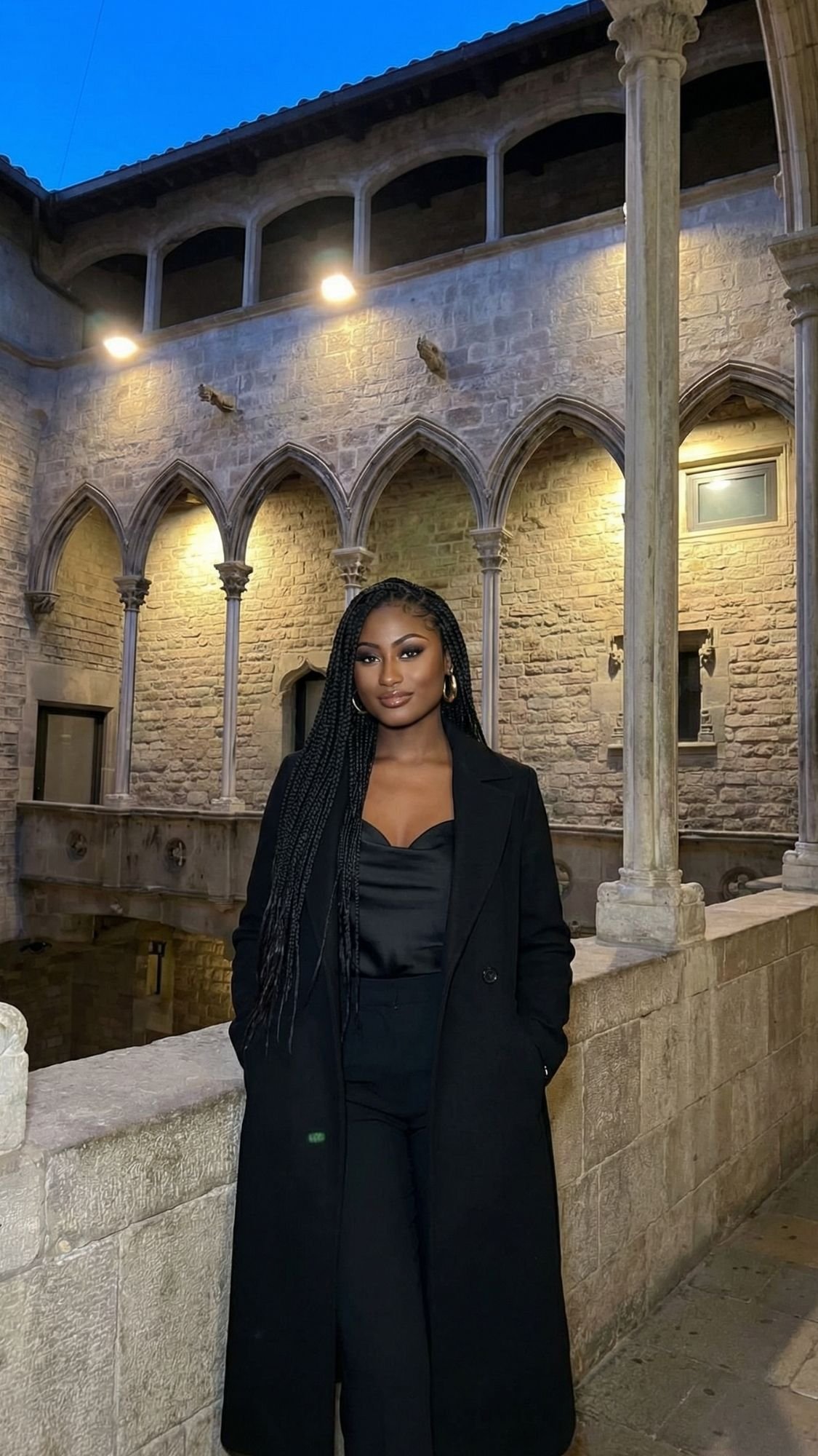 A woman with long braids, dressed in a black coat and outfit, stands in front of a historic stone building with arches and columns, illuminated by warm lights—a perfect scene for winter plans in Barcelona.
