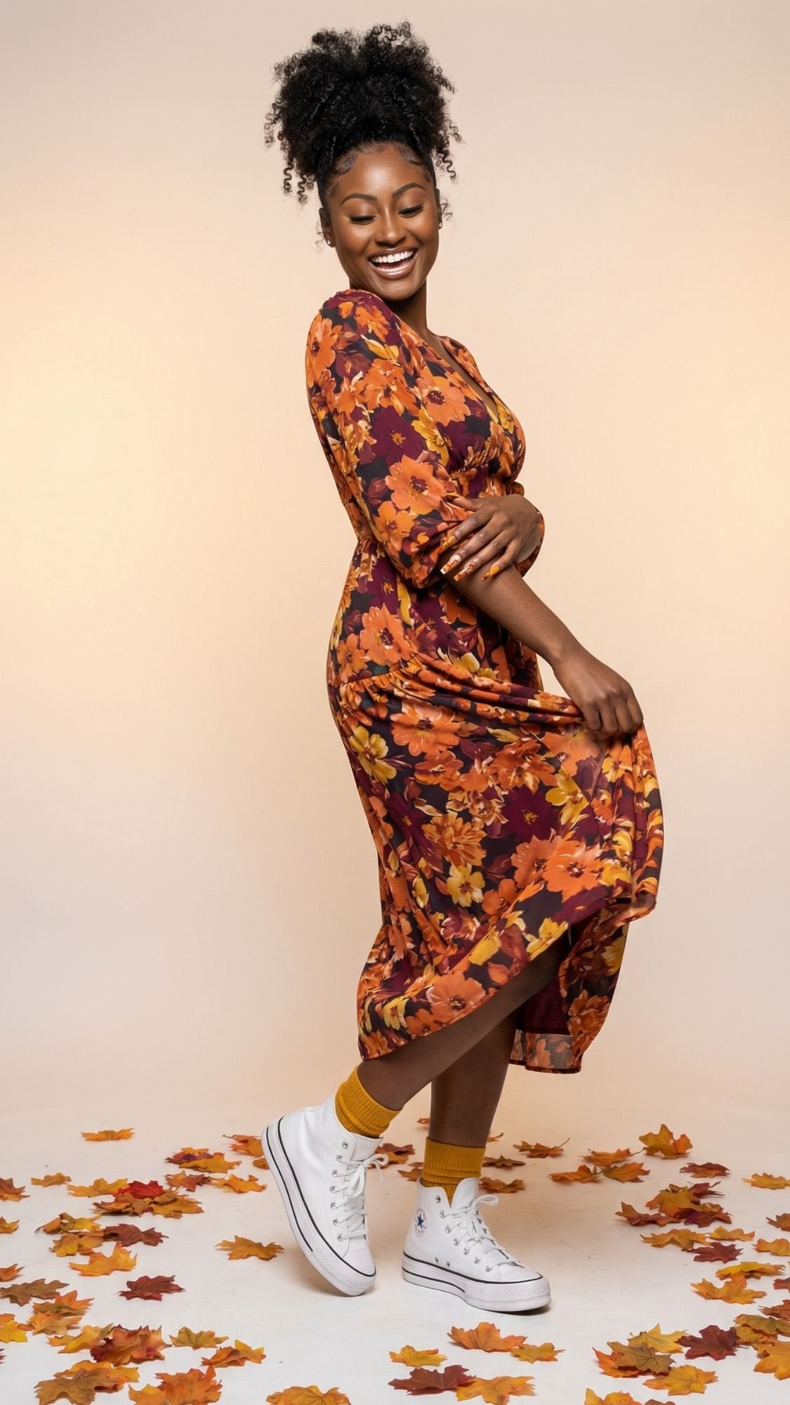 A smiling woman in a fall-themed floral dress and white sneakers poses playfully, surrounded by scattered autumn leaves—showcasing dopamine dressing against a light beige background.