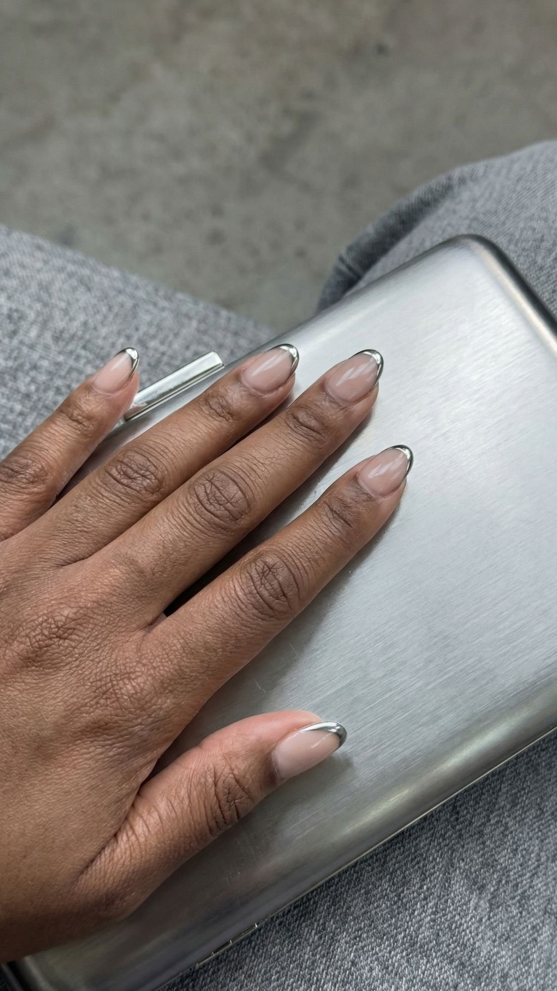 A hand with neatly manicured French tip nails featuring a natural base and metallic silver tips rests on a silver metallic clutch, exuding quiet luxury. The background includes gray fabric and a concrete surface.