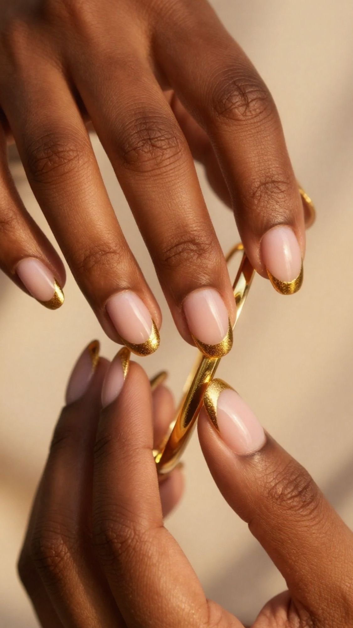 Close-up of hands with natural almond-shaped French tip nails featuring shiny gold tips and quiet luxury, holding a simple gold bangle against a soft neutral background.