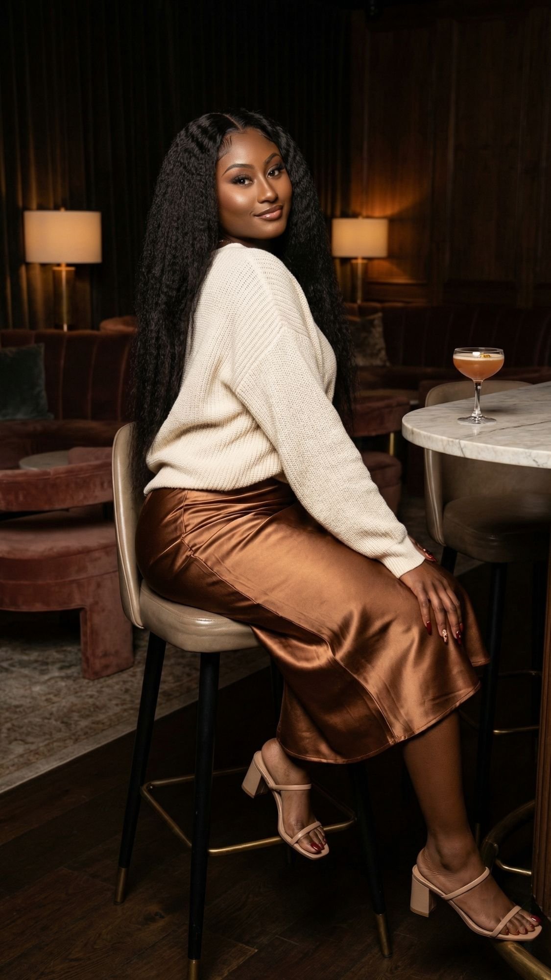 A woman with long, curly hair sits on a bar stool in a stylish lounge, embracing fall fashion in a cream sweater, bronze satin skirt, and beige sandals. She smiles slightly, with a cocktail glass on the marble table beside her.