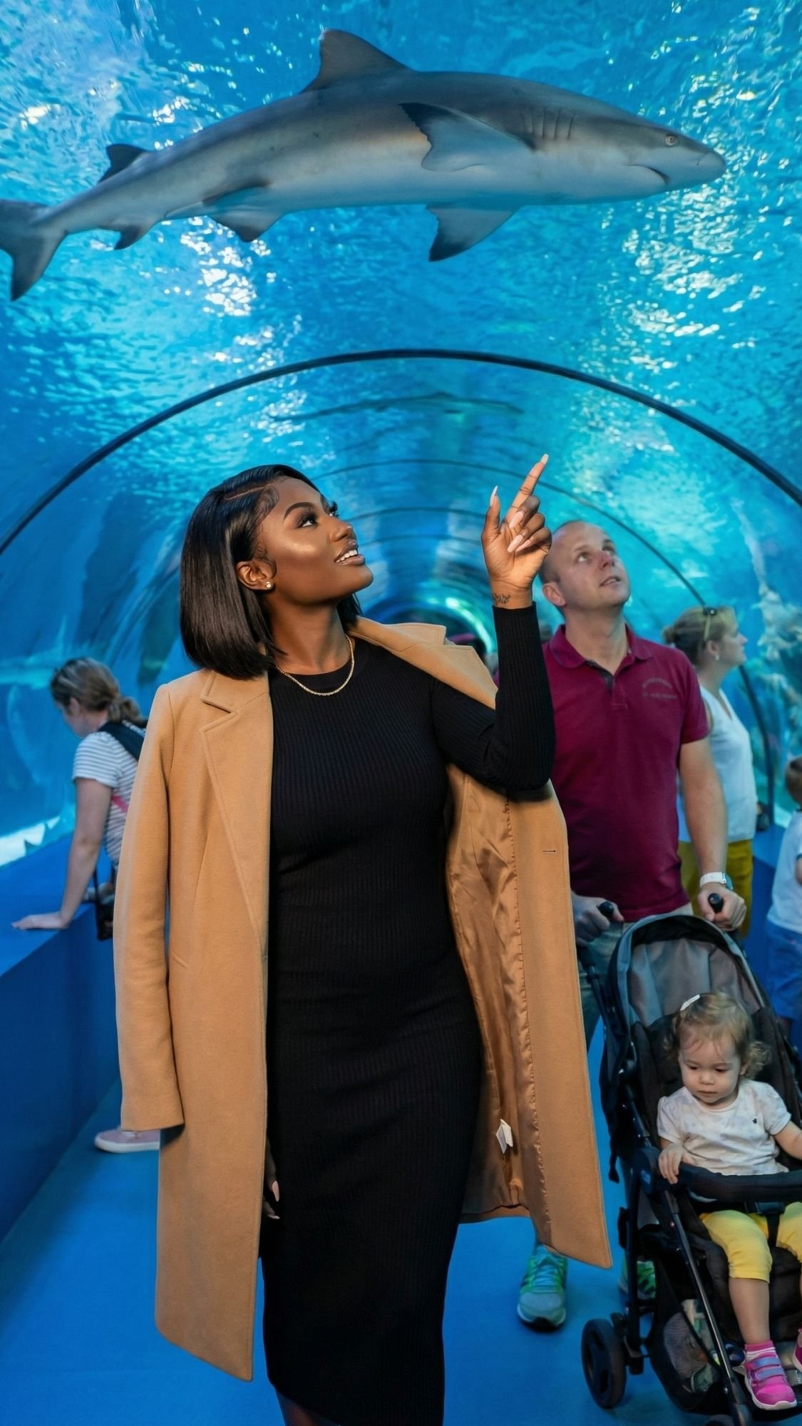 A woman in a tan coat points up at a swimming shark inside an aquarium tunnel. Other visitors, including a man with a stroller, admire the marine life above—a perfect off season Barcelona activity beneath blue water and swirling fish.