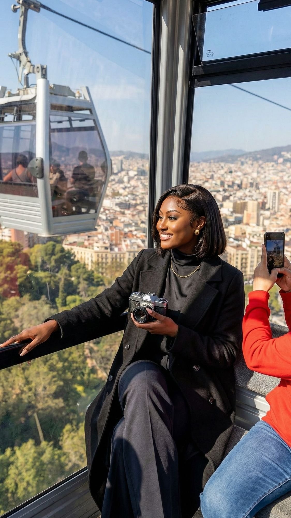 A woman in a black coat smiles and holds a camera while riding a cable car over Off Season Barcelona. Another person next to her takes a photo with a phone, capturing the city and green hills visible through the window.