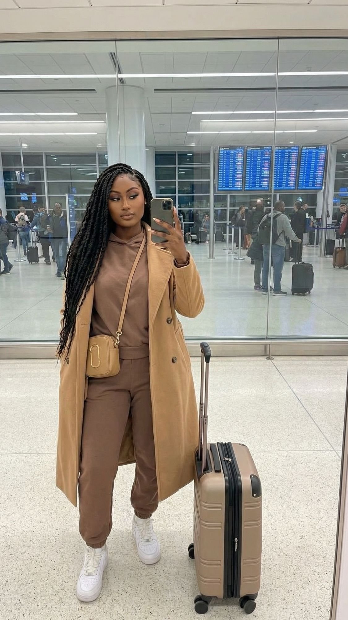 A woman in a tan coat, brown outfit, and white sneakers stands in an airport with a matching suitcase, snapping a mirror selfie. The scene captures quiet girl winter outfits and calm mornings amidst travelers and flight info screens.
