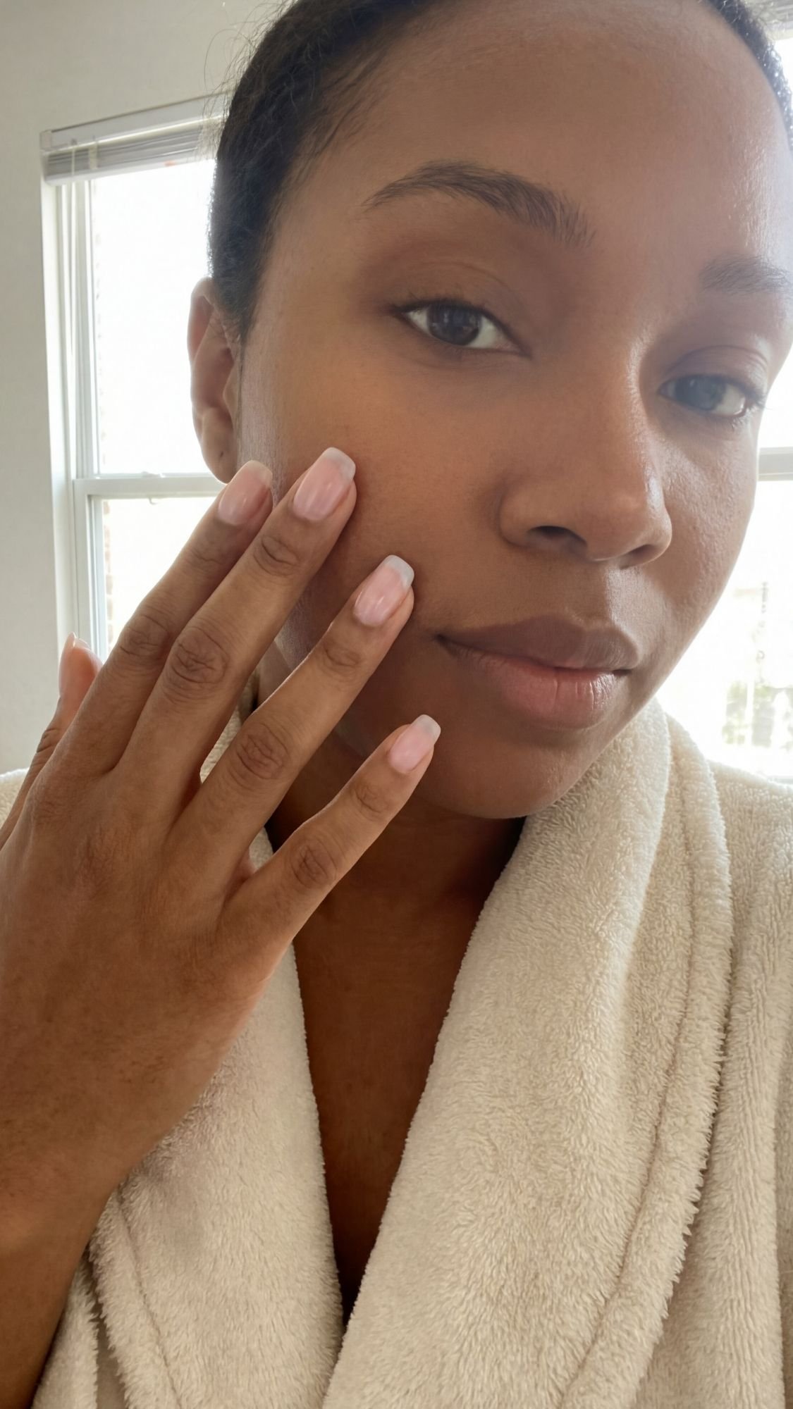 A woman in a white bathrobe touches her face gently, showing clear skin and elegant French tip nails. She has a calm expression and stands in front of a window with natural light, capturing the essence of quiet luxury.