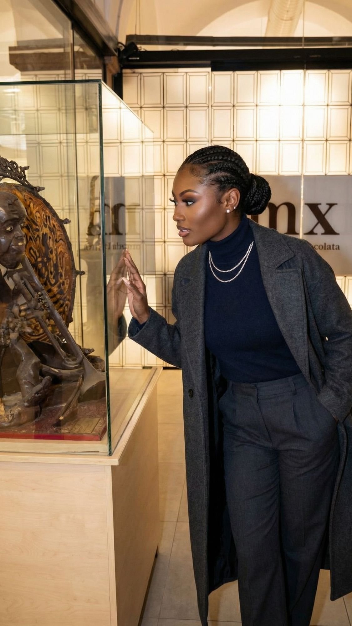 A woman in a dark coat and turtleneck admires a detailed statue inside a glass display case at a museum—embracing cozy winter plans during off season Barcelona, she leans in closely with interest, one hand gently touching the case.