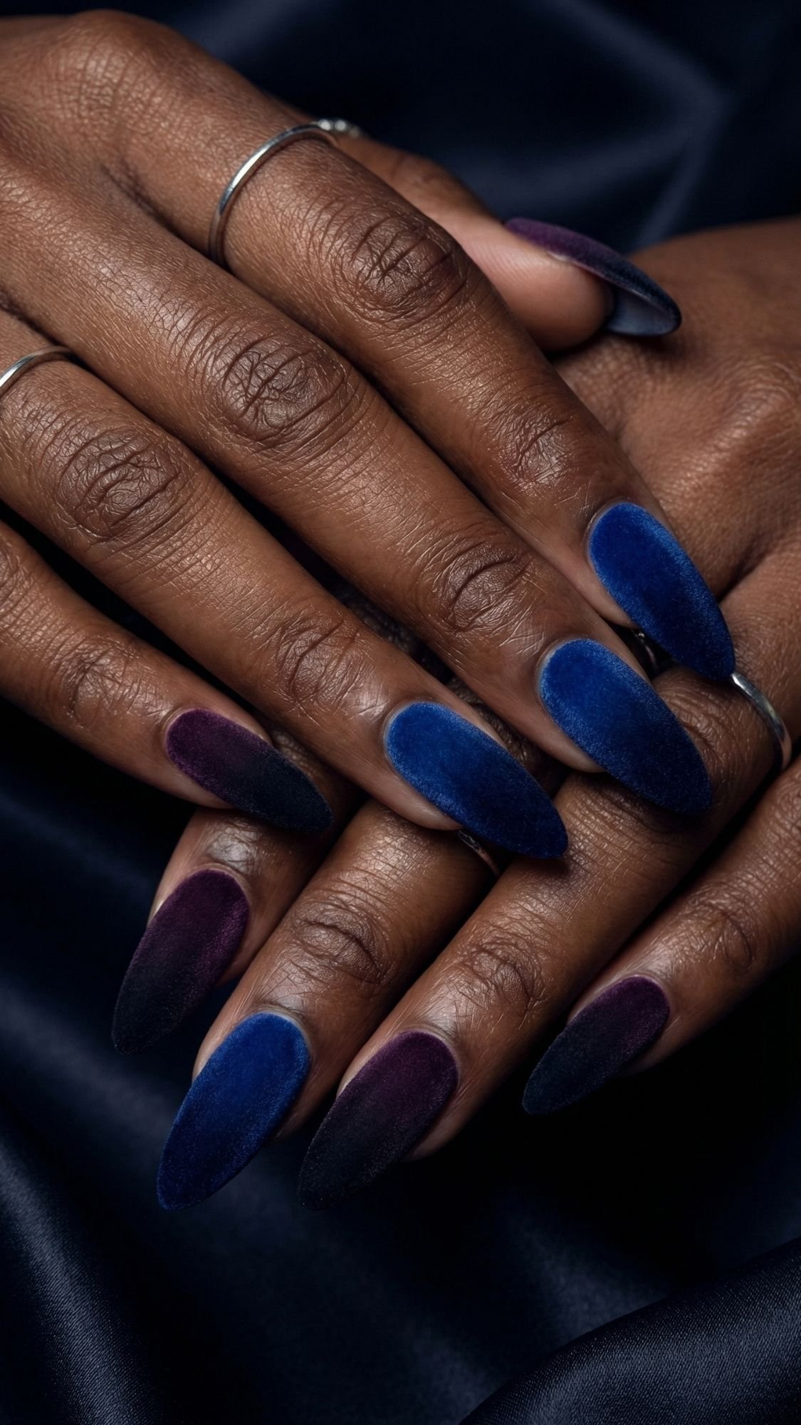 A close-up of elegant nails with an almond shape and gradient velvet finish in deep blue and purple shades, perfect for holiday parties. The hands, adorned with thin silver rings, rest gracefully on dark fabric.