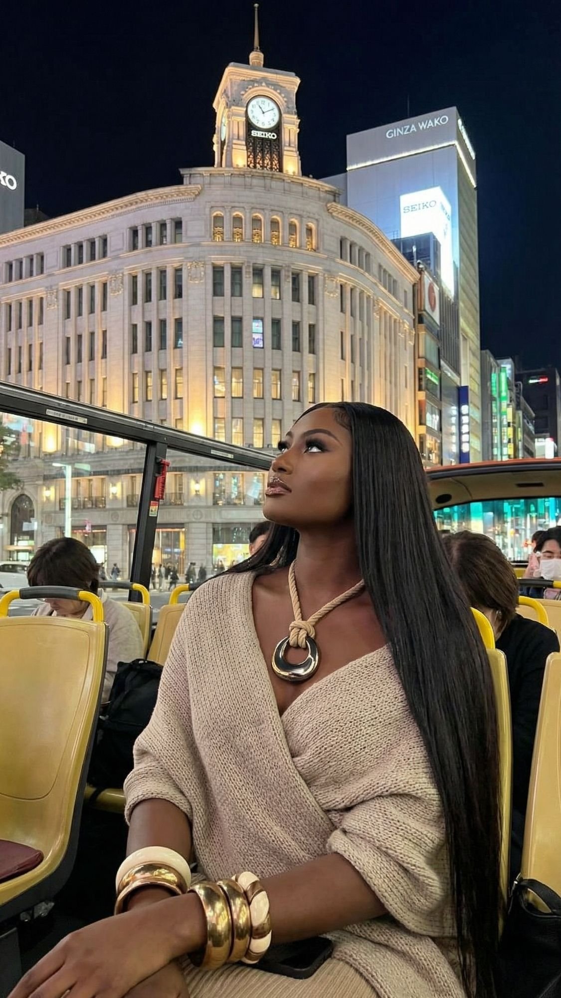 A stylish woman with long straight hair sits on a city tour bus at night, wearing a beige outfit and bold jewelry. Illuminated buildings and a clock tower glow in the background—perfect for exploring Tokyo Winter Illuminations with other passengers nearby.