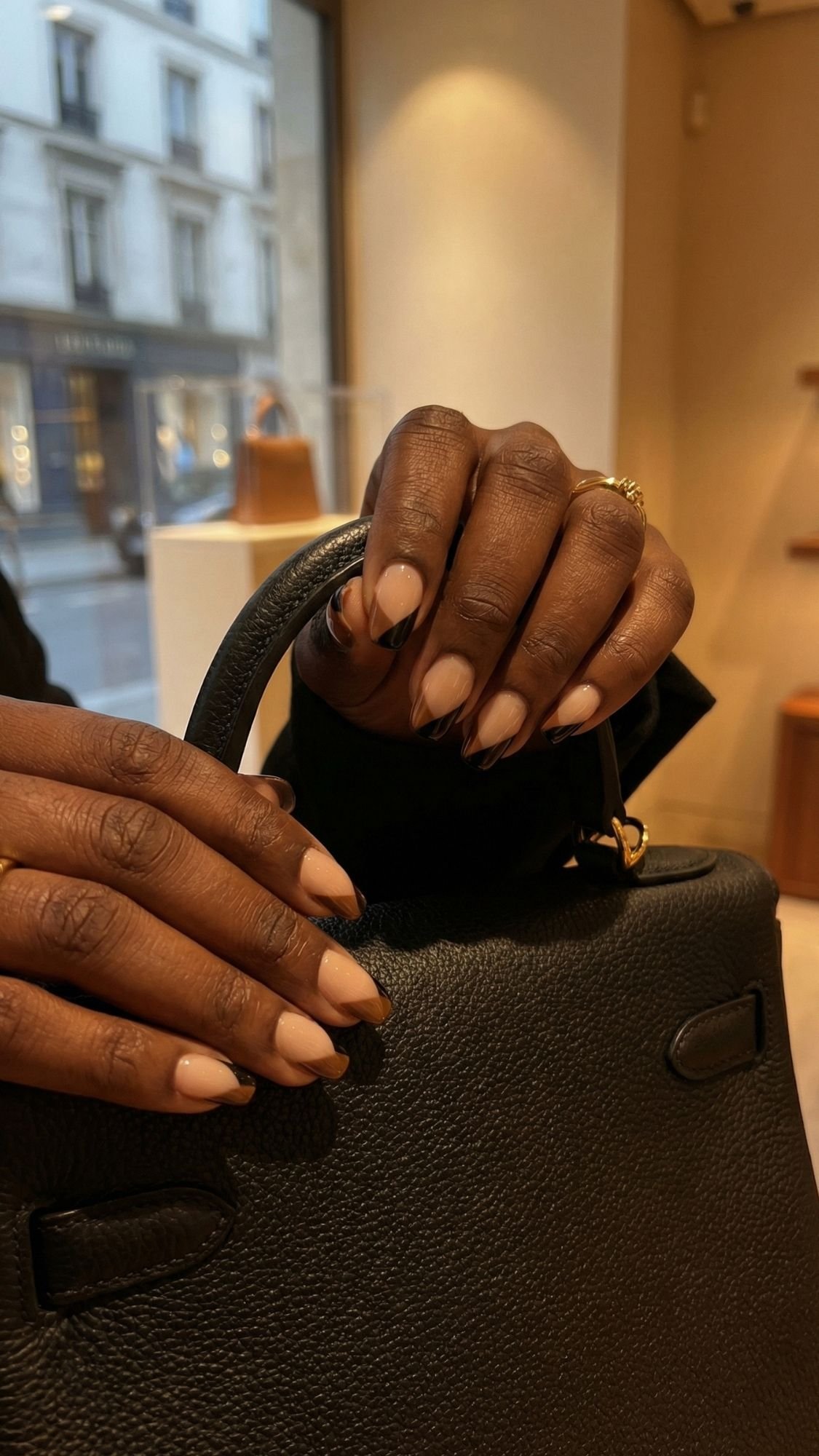 A close-up of hands with neatly manicured French tip nails holding the handle of a black leather handbag inside a store, channeling quiet luxury, with a street scene visible through the window in the background.