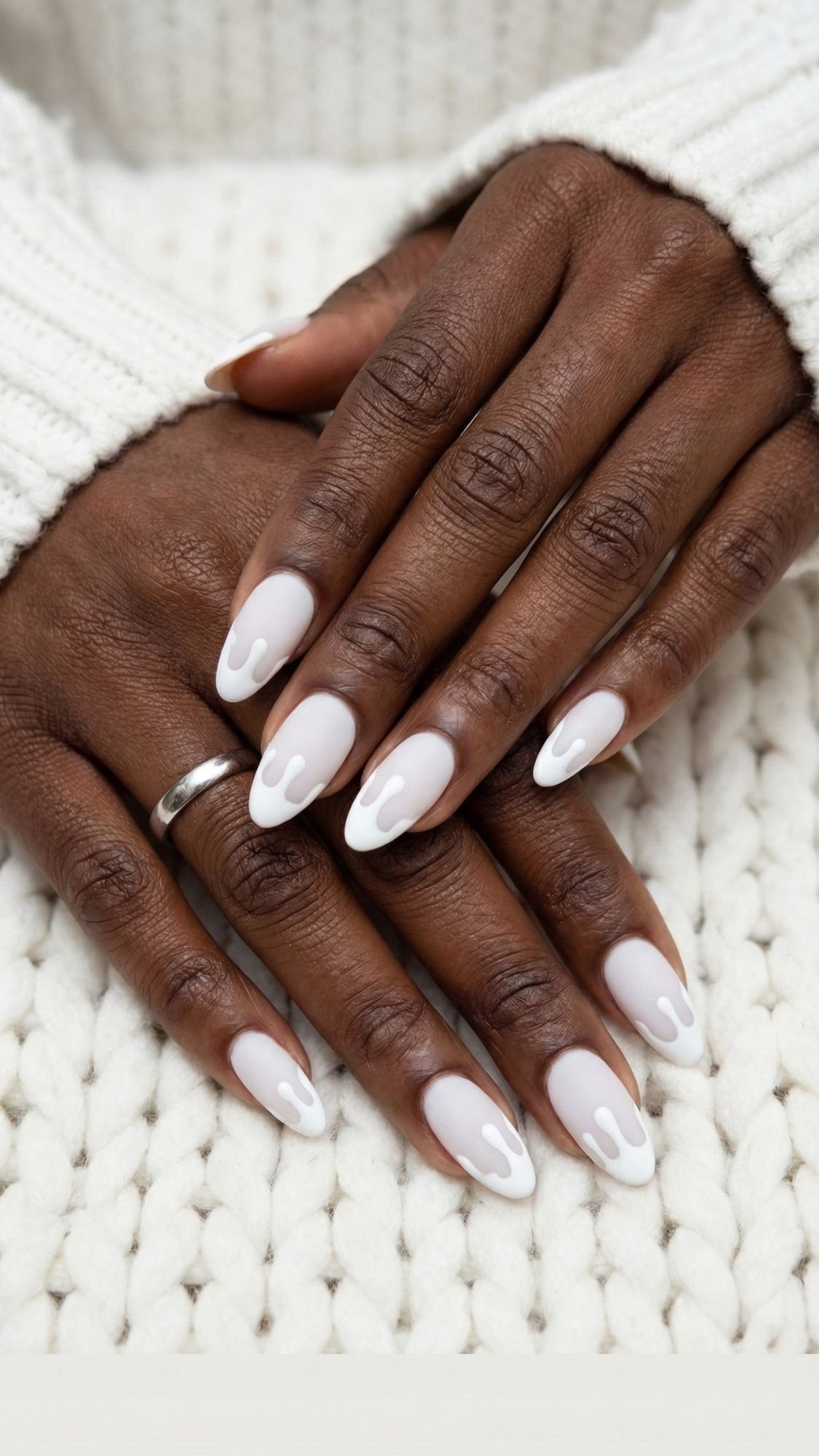 Close-up of hands with elegant, almond-shaped nails in a white and light pink drip design—perfect for holiday parties. A silver ring and chunky white knit sweater complete this chic look, with hands resting on a matching knit surface.