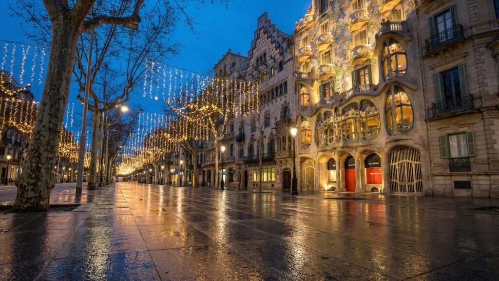 A wet city street at dusk in off season Barcelona glows with festive lights above, their reflections shimmering on the pavement. Ornate historic buildings, including one with a wavy facade, are warmly lit as bare trees line the quiet avenue.