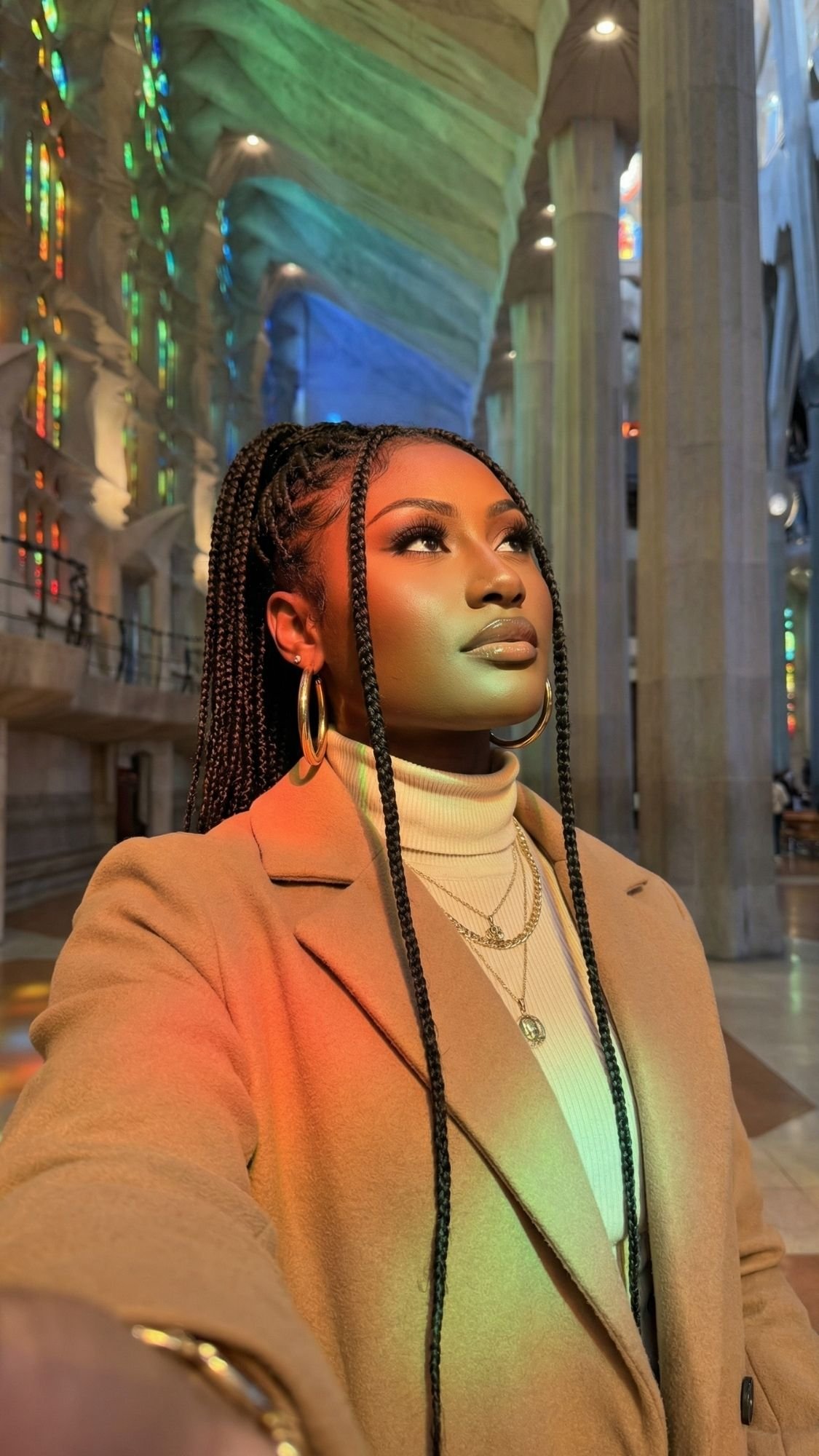 A woman with long braids, wearing a camel coat and white turtleneck, takes a selfie inside a grand building—one of the cozy things to do in Barcelona during winter—its tall columns and stained glass casting vibrant light around her.