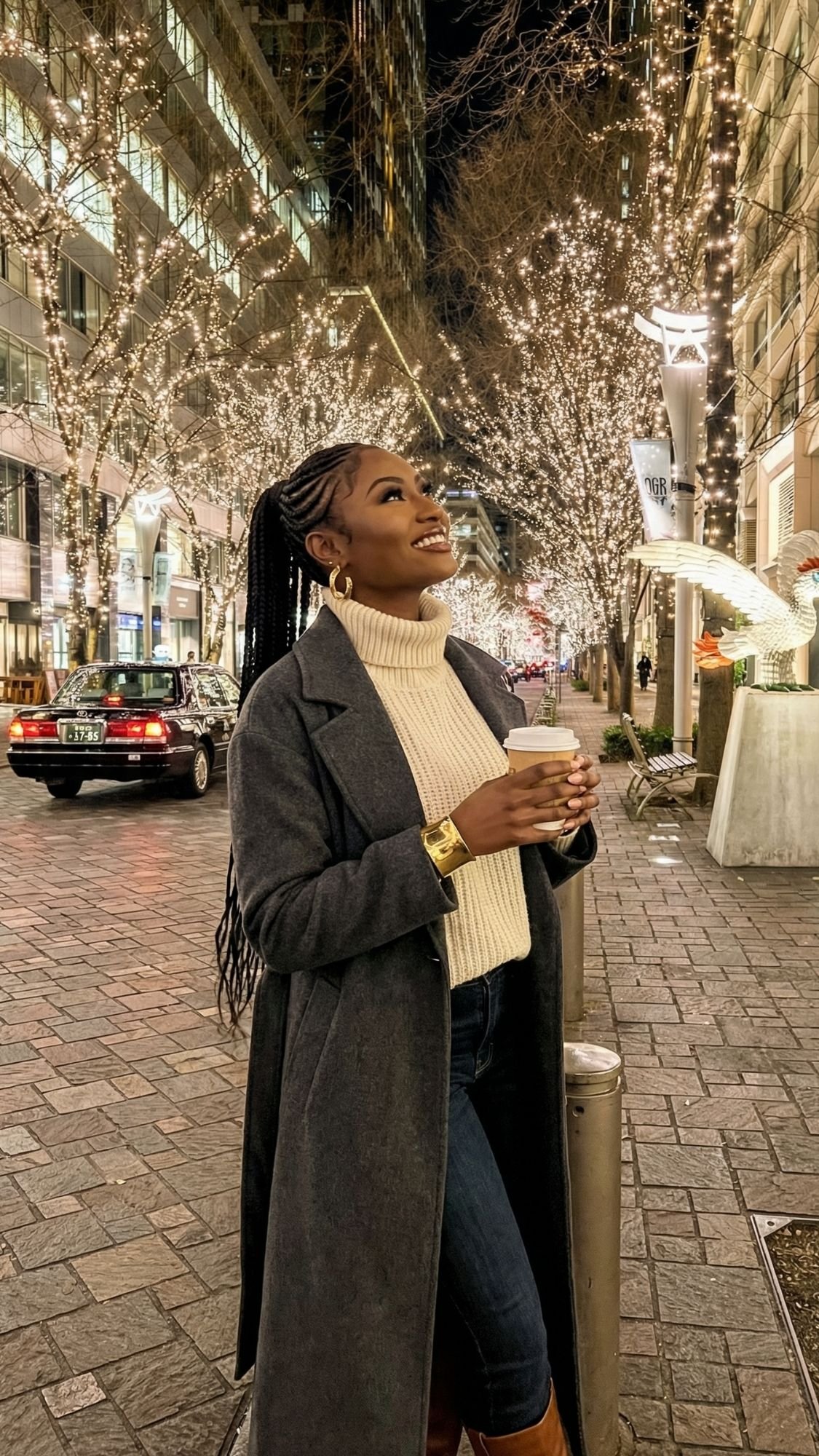 A woman in a gray coat and white turtleneck holds a coffee cup and smiles, looking up at festive Tokyo winter illuminations on trees lining a city street at night. Cars and illuminated buildings glow in the background.