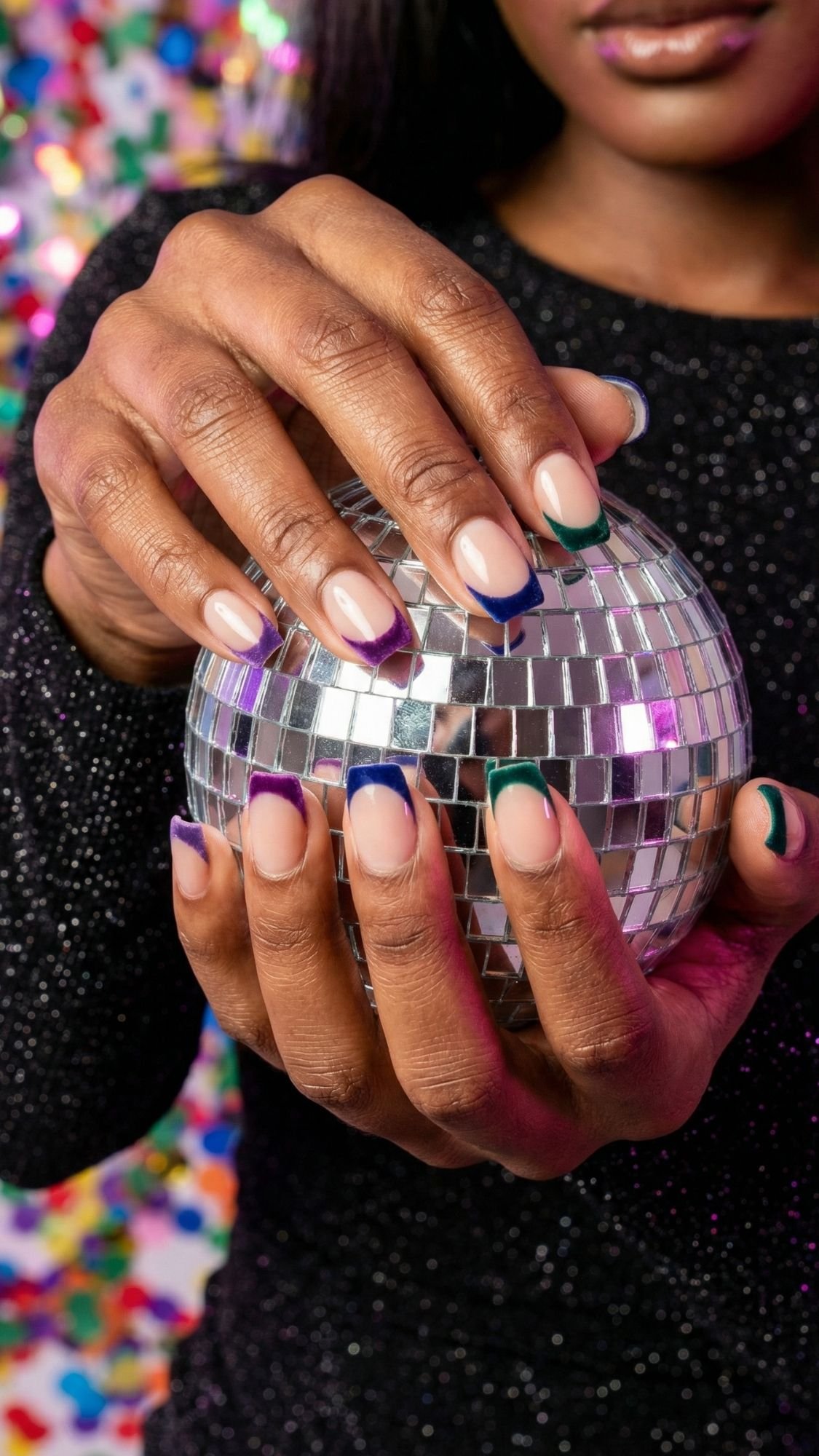 A person with neatly manicured, elegant nails featuring colorful French tips holds a small disco ball. The festive background sparkles with blurred confetti, perfect for holiday parties, while the person wears a chic, black glittery top.