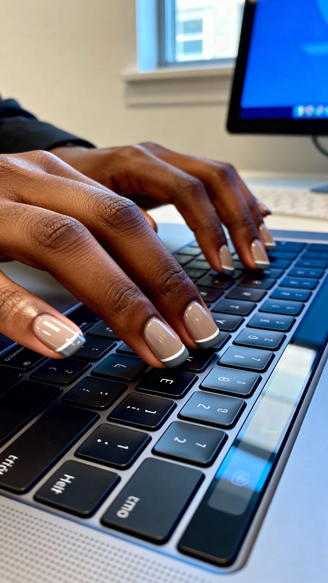 Close-up of hands with fall nails typing on a laptop keyboard. A desktop monitor is visible in the background, with sunlight streaming through the window, capturing an air of quiet luxury.