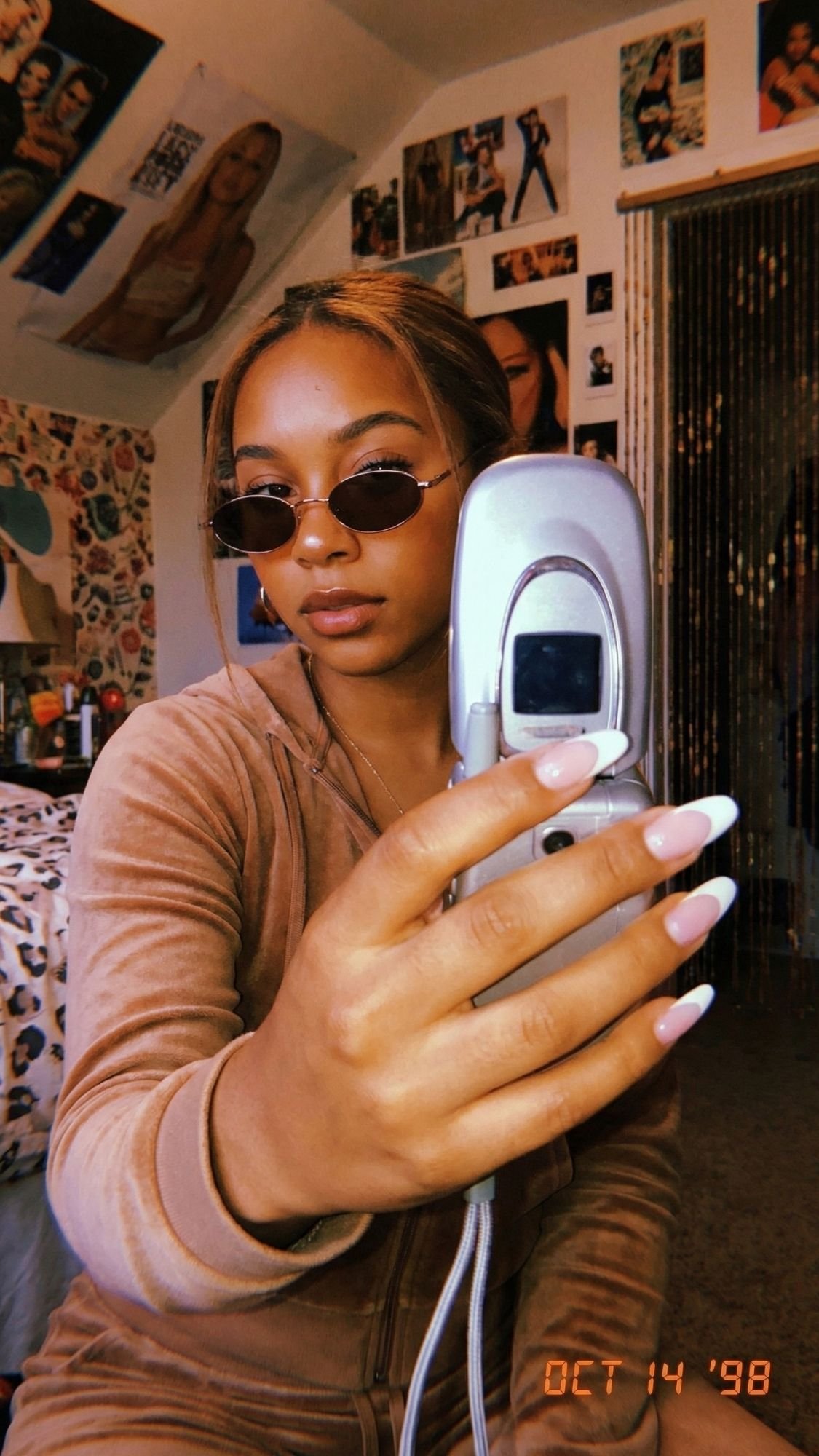 A woman in a velour tracksuit and sunglasses takes a selfie with a flip phone in a bedroom decorated with posters, showing off her French tip nails. The date Oct 14 98 is displayed in the corner.