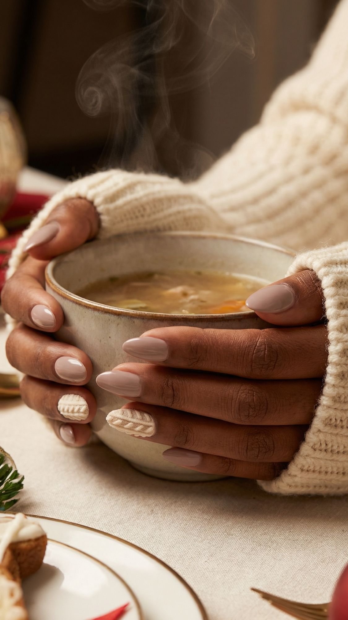 A person wearing a cream-colored sweater holds a steaming bowl of soup with both hands. Their nails are manicured in soft beige tones with a sweater knit design on two accent nails—perfect elegant nails for holiday parties.