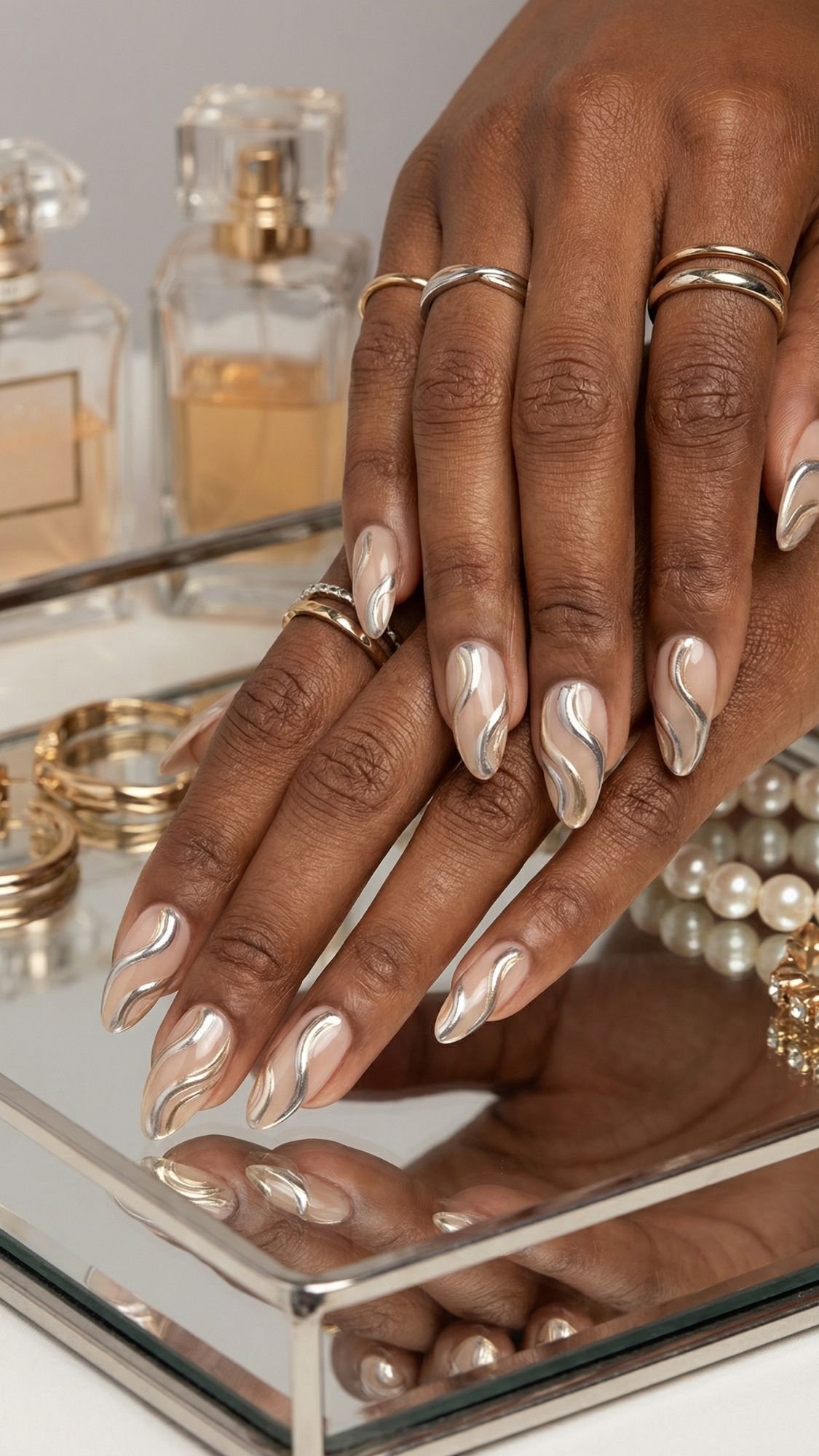 A close-up of hands with elegant nails featuring shiny, nude and metallic swirl art, wearing multiple gold rings, resting on a mirrored tray with perfume bottles, pearl jewelry, and gold accessories—perfect chic ideas for holiday parties.