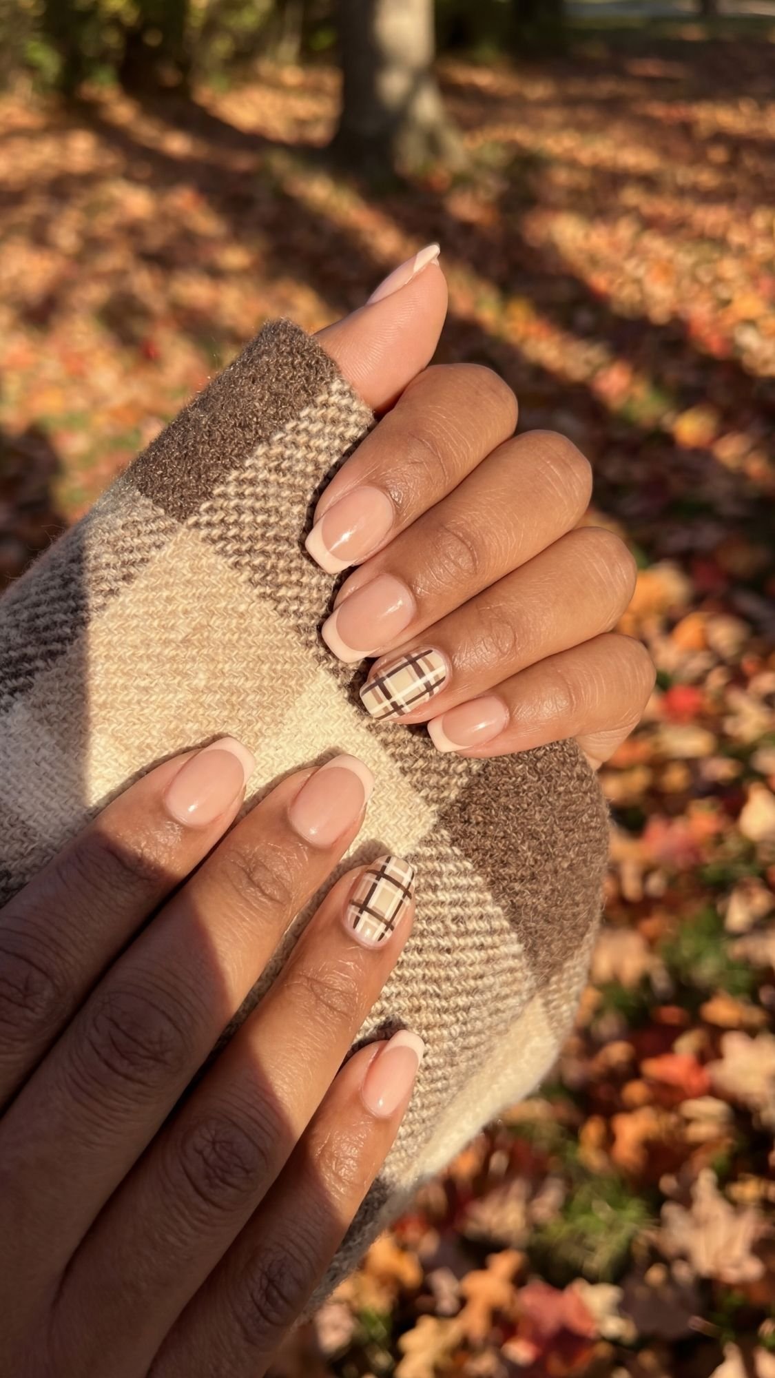 A person with medium-brown skin shows neatly manicured fall nails painted nude, two accent nails in a plaid design. Wearing a brown plaid coat, they embody quiet luxury with autumn leaves scattered on the ground in the background.