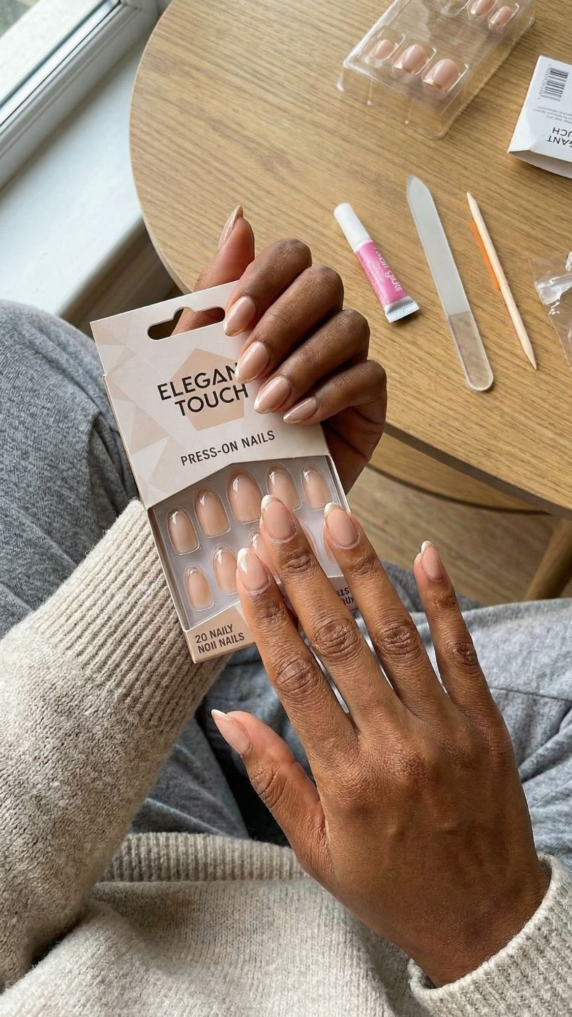 A person with medium brown skin is applying nude press-on nails from an Elegant Touch kit at a wooden table—perfect for chic ideas and elegant nails that will stand out at holiday parties. Nail tools, glue, a nail file, and extras are also on the table.