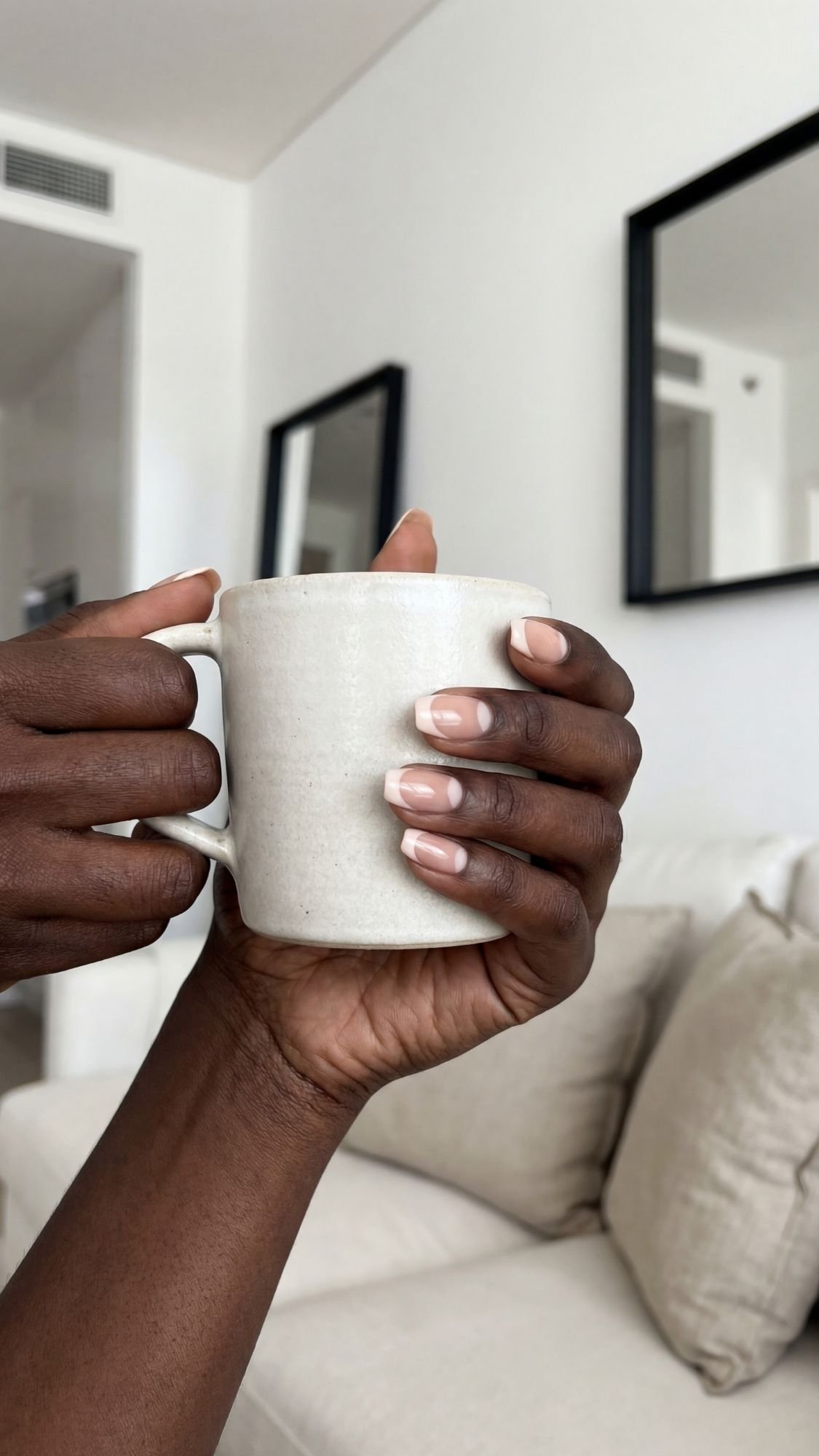 Close-up of hands with chic French tip nails holding a white ceramic mug in a modern, minimalistic living room with neutral-colored sofa, pillows, and two black-framed mirrors—a perfect scene of quiet luxury.