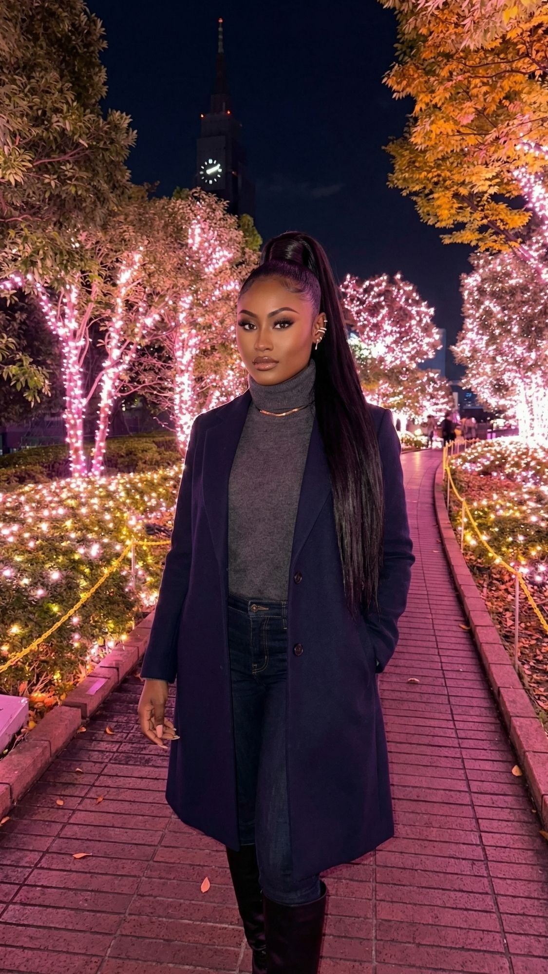 A woman wearing a dark coat and turtleneck stands on a brick path lined with trees decorated in bright pink lights at night, capturing the magic of Tokyo winter illuminations. Tall buildings and more glowing trees light up the background.