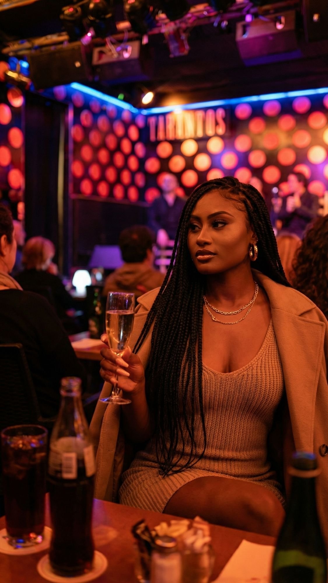 A woman in a tan outfit sits at a table in a dimly lit nightclub, embracing cozy winter plans. She holds a glass of champagne, her long braided hair and gold jewelry shining as musicians perform on a stage with colorful circular lights.