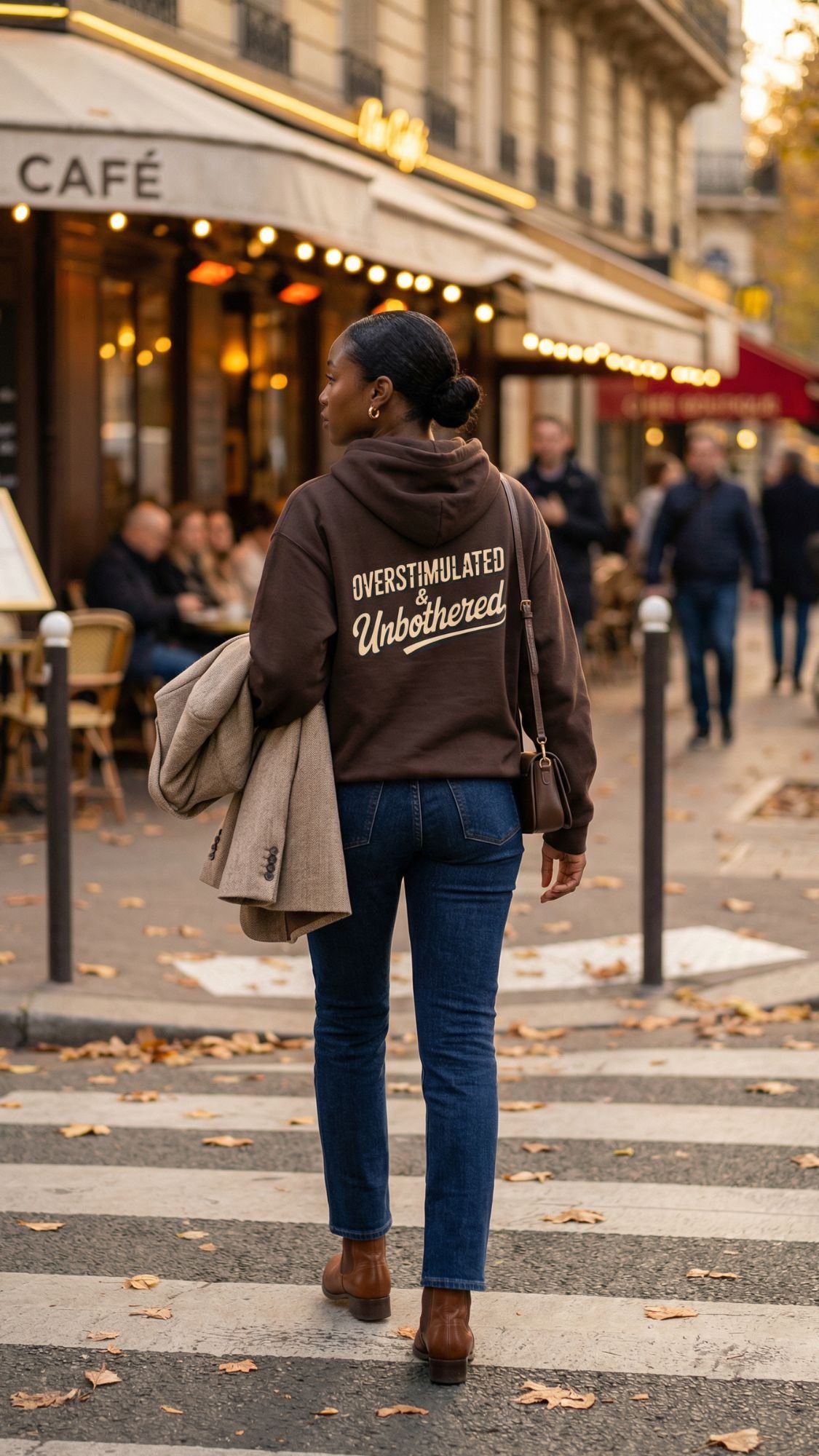 A person walks on a city street crosswalk in jeans, brown boots, and a back print hoodie that says Overstimulated but Unbothered—perfect for fall travel and inspiring hoodie outfit ideas—with a café and people in the background.