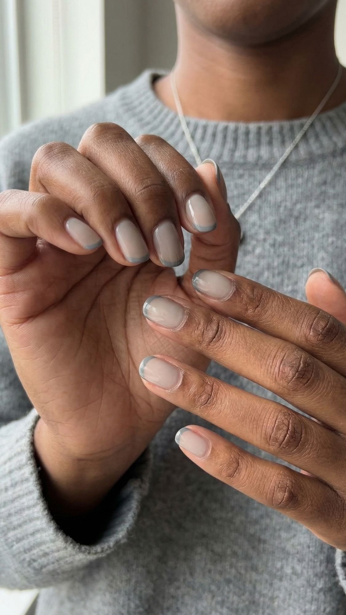 A person wearing a gray sweater shows off their hands with short, neatly manicured French tip nails featuring a light nude polish and thin silver tips, embracing the quiet luxury and subtle elegance of fall vibes.