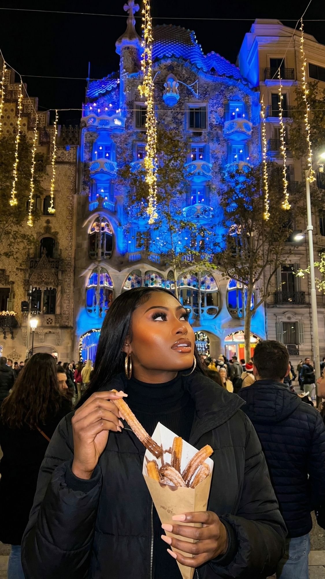 A woman in a black jacket holds churros at night in front of a beautifully illuminated building with blue and golden string lights. People are gathered, enjoying the festive atmosphere—perfect for cozy winter Barcelona adventures.