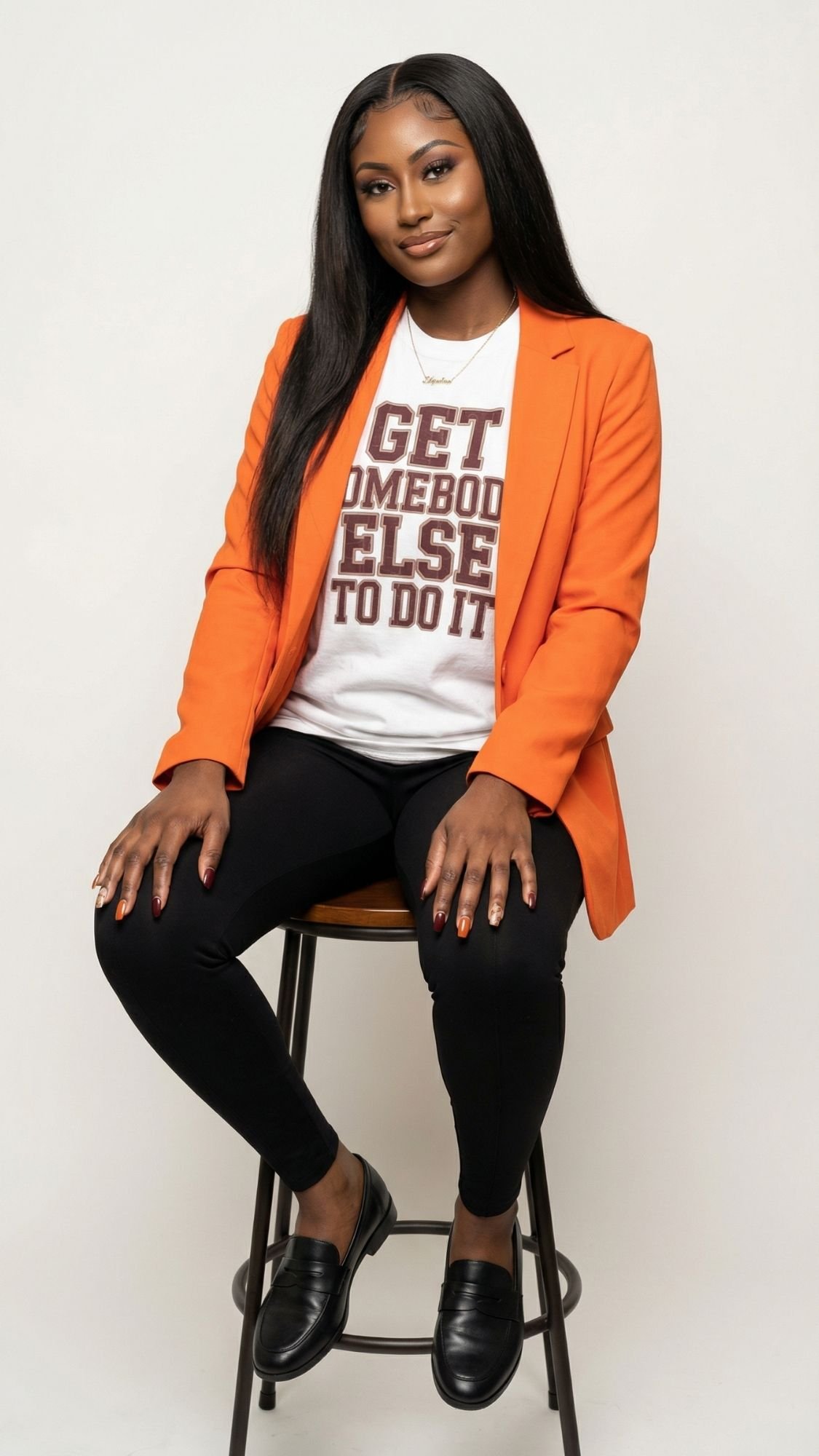 A woman with long straight hair sits on a stool, wearing a bright orange blazer for some dopamine dressing, a white t-shirt with GET SOMEBODY ELSE TO DO IT in bold letters, black pants, and black loafers, against a plain backdrop.