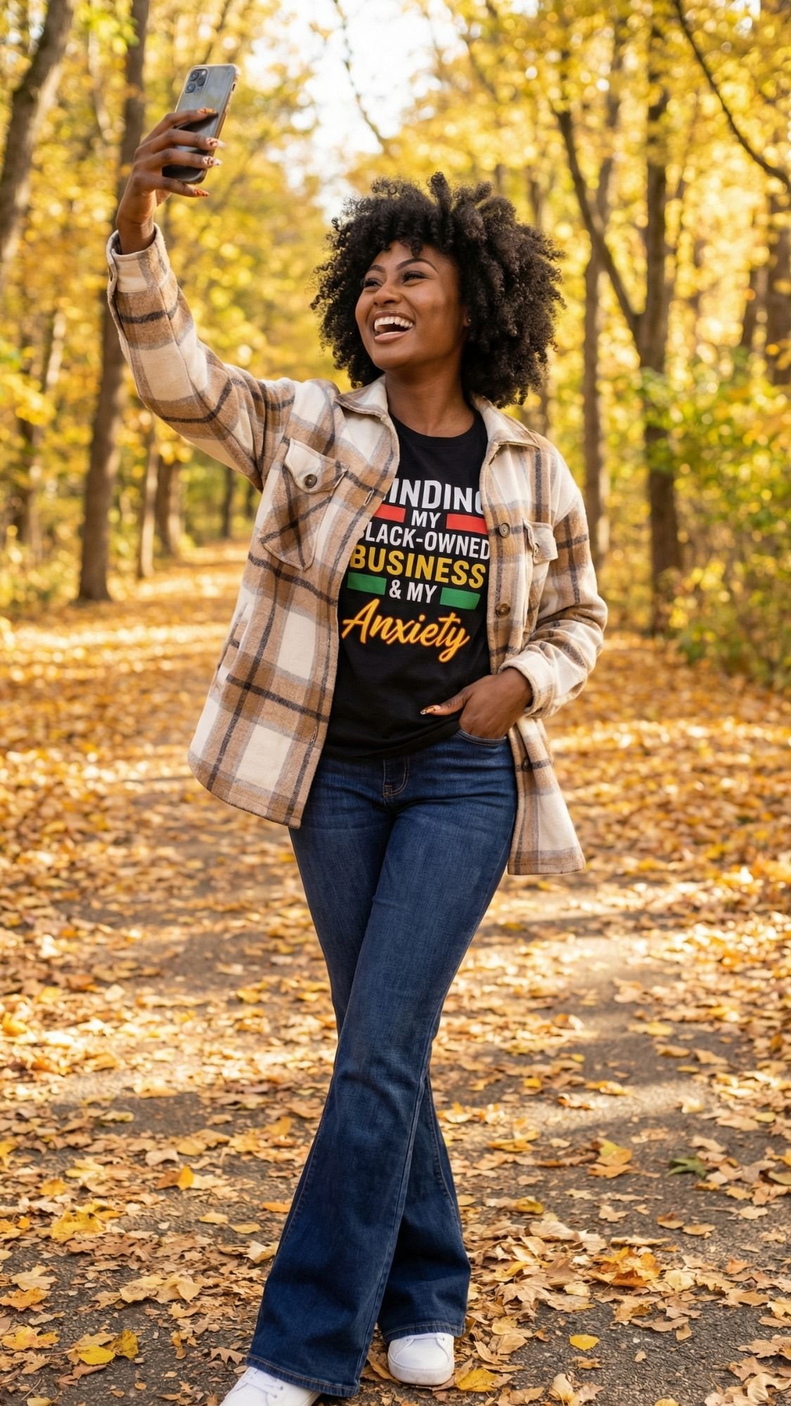 A woman stands on a leaf-covered path in a sunlit forest, smiling and taking a selfie. She wears fall fashion—a plaid jacket, jeans, and a Black-owned business T-shirt—embracing dopamine dressing with style and confidence.