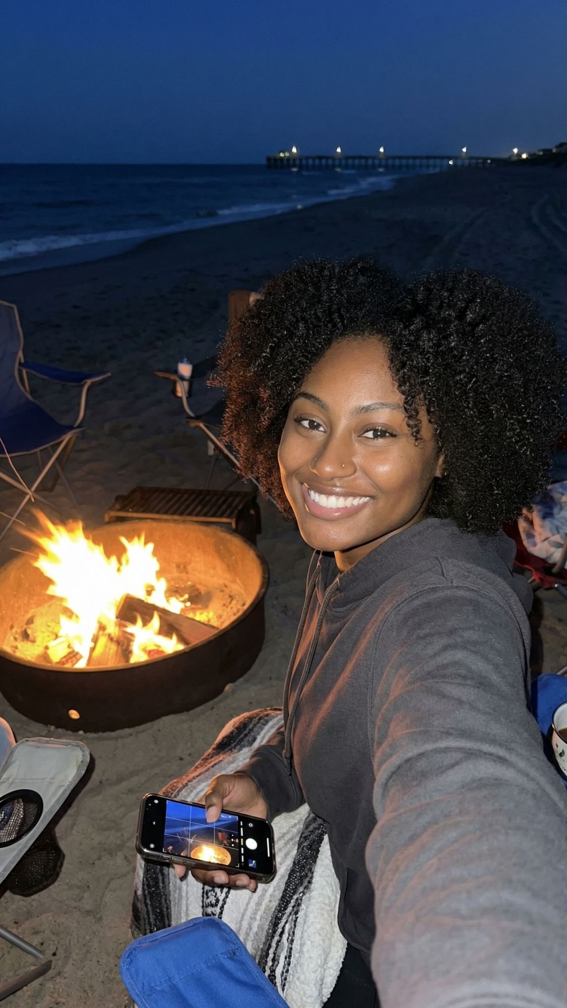 A woman takes a selfie while smiling at a beach campfire during her Fall Getaway. She sits wrapped in a blanket under the dark sky, with waves and an Outer Banks pier visible in the background, soaking up the coastal vibes.