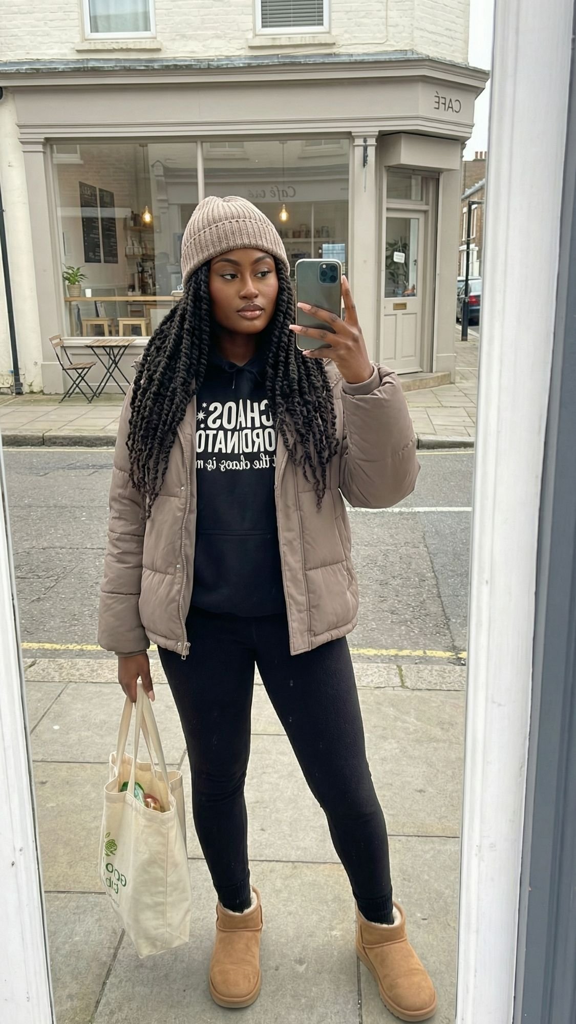 A woman takes a mirror selfie on a sidewalk, showcasing her Quiet Girl Winter Outfit with a beige beanie, tan puffer jacket, black hoodie, and tan boots. Across the street, a café sets the scene for calm mornings.