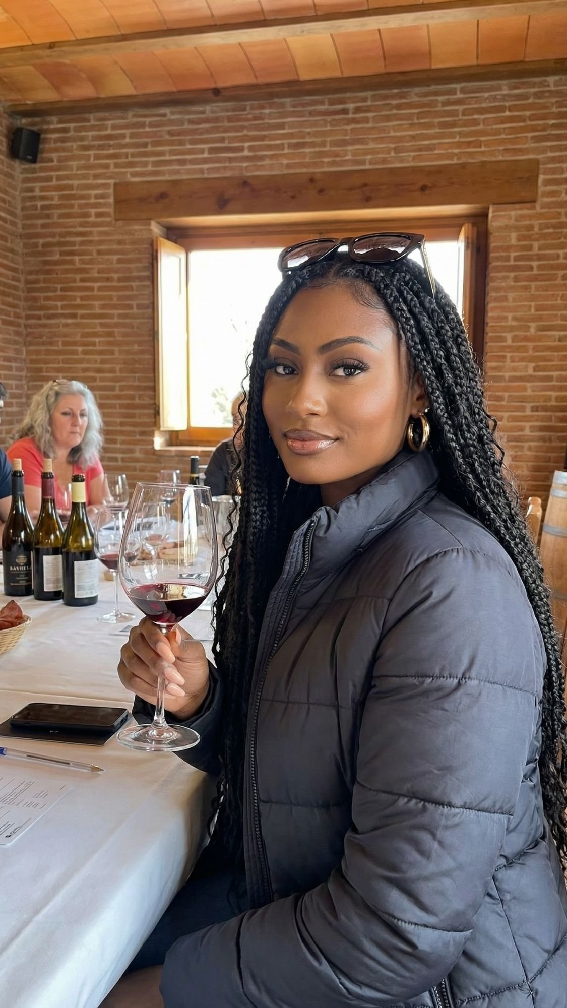 A woman with long braids and sunglasses on her head sits at a table, holding a glass of red wine. She wears a dark jacket. Behind her, others enjoy bottles of wine in a brick-walled room—perfect for cozy winter plans in Barcelona.