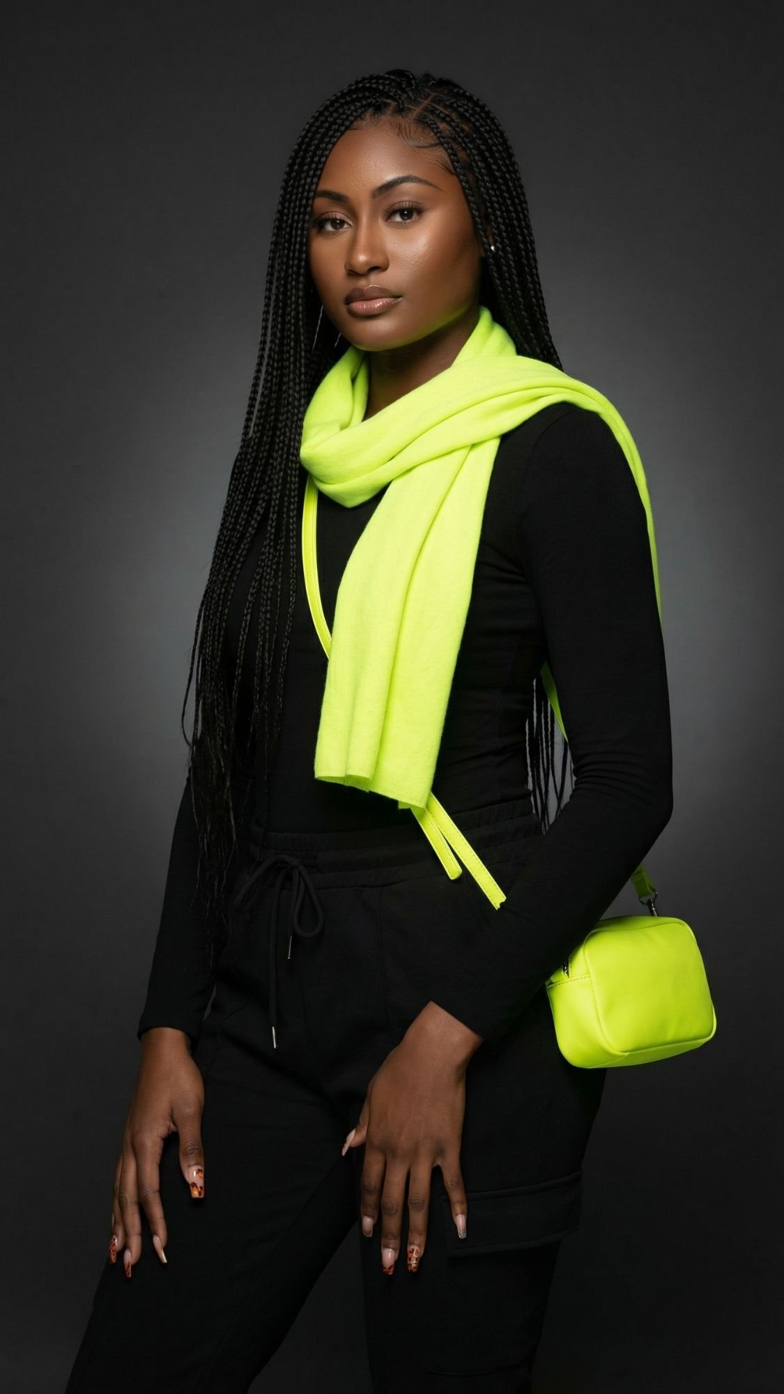 A woman with long braids wears a black outfit paired with a neon yellow scarf and matching small purse, embracing dopamine dressing as she poses against a dark background.