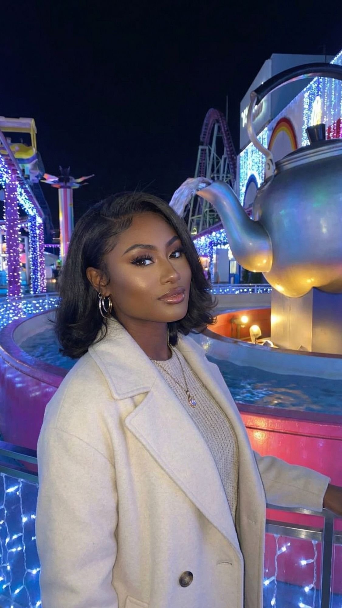 A woman with shoulder-length black hair and a beige coat poses in front of a large teapot sculpture and colorful lights at a Tokyo Winter Illuminations event, capturing the magic of nighttime amusement park adventures.