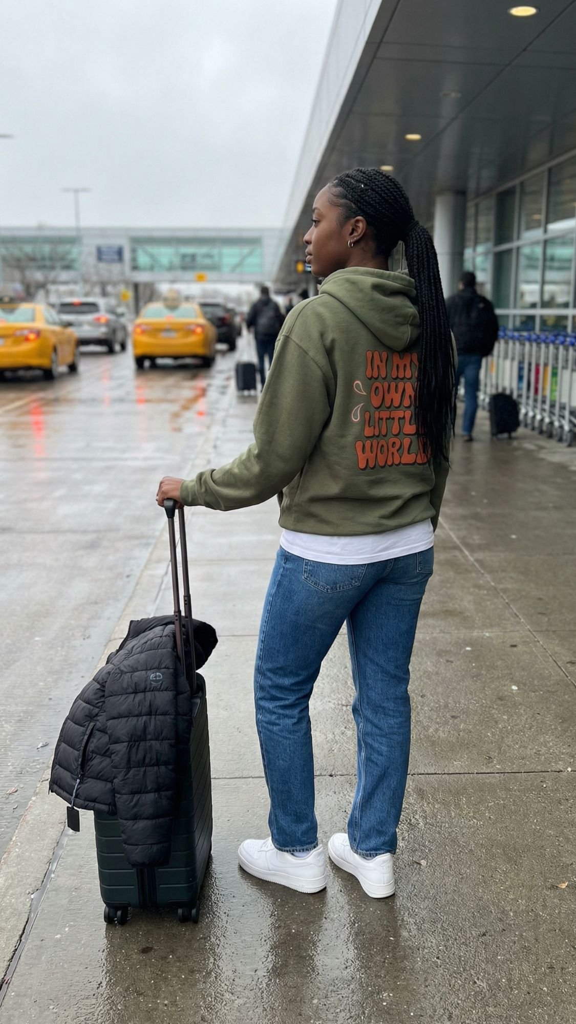 A person stands on a wet sidewalk outside an airport in a stylish fall travel look, wearing jeans, white sneakers, and a green back print hoodie that says IN MY OWN LITTLE WORLD. Yellow taxis are visible in the background.