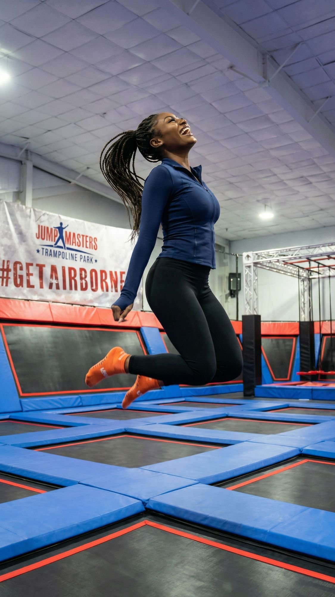A woman in athletic clothing smiles mid-air, jumping on blue and black trampolines at an indoor trampoline park with #GETAIRBORNE and Jump Masters banners—embracing those fun Coastal Vibes perfect for a Fall Getaway near the Outer Banks.