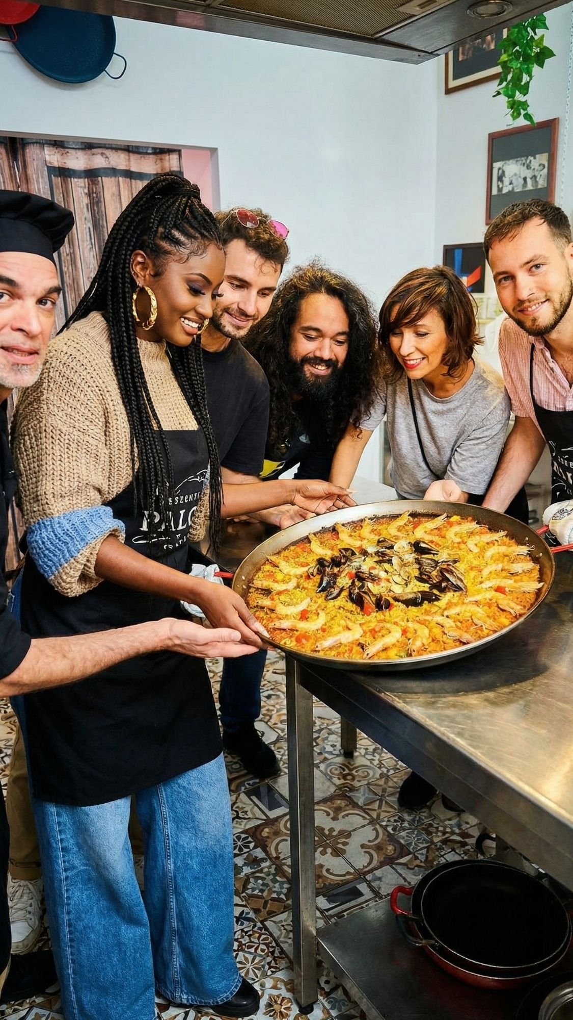A group of six people, smiling and gathered around a large pan of paella on a kitchen counter, pose together in a cozy kitchen setting—perfect for making Cozy Plans during winter in Barcelona.