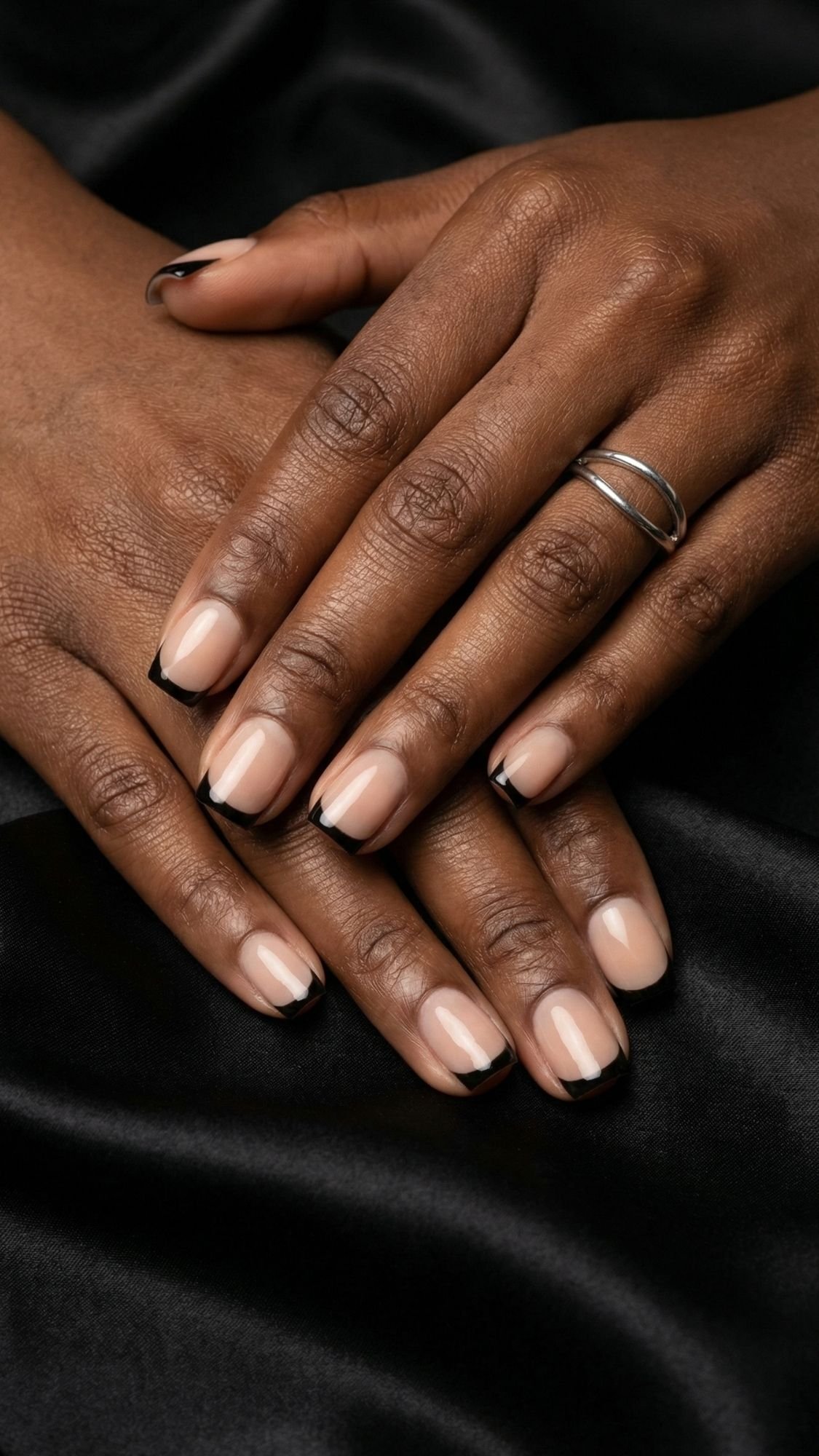 Two hands with medium-dark skin rest on black satin fabric. The nails have a nude base with thin black French tips—an elegant nails look, perfect for chic ideas at holiday parties. One hand wears a simple silver double-band ring.