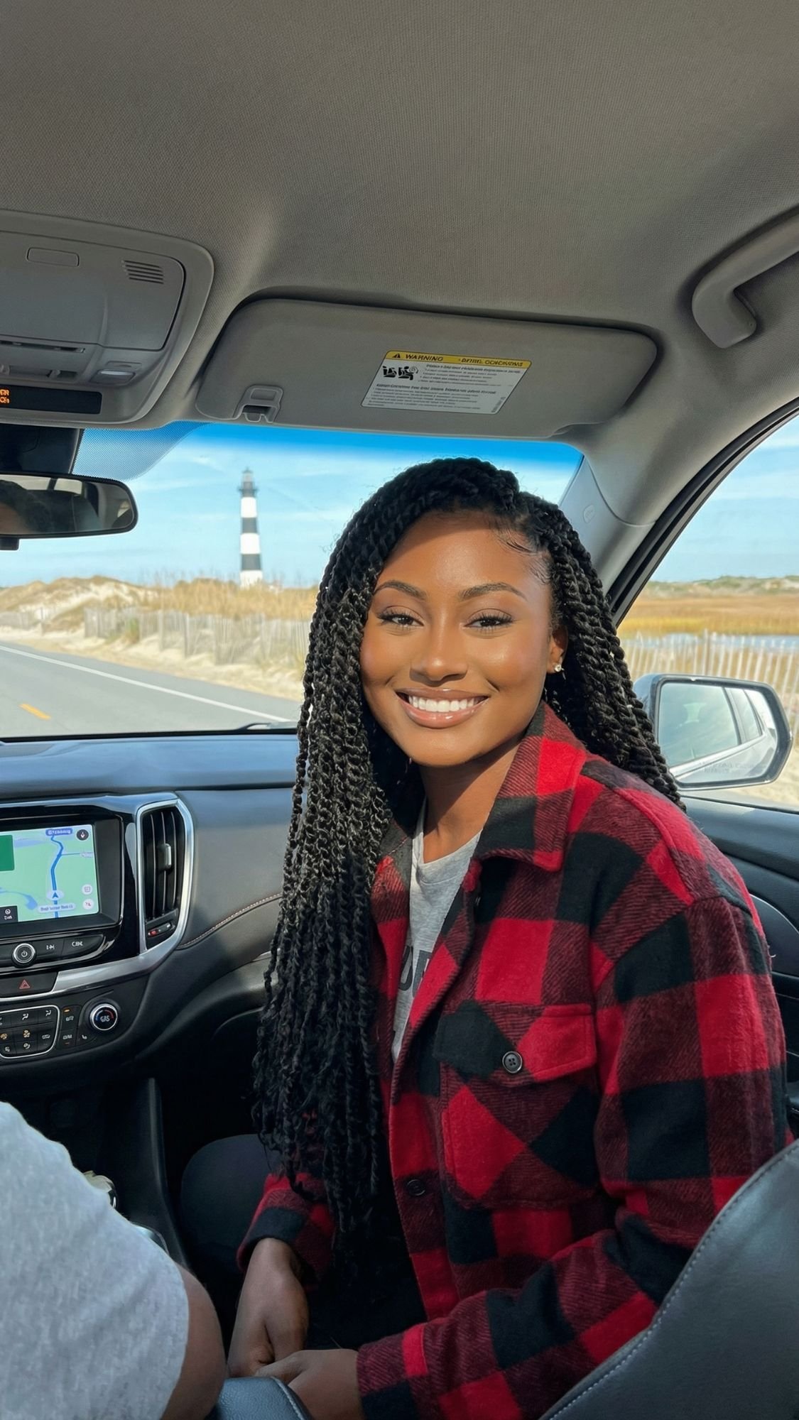 A smiling woman with long braids and a red plaid jacket sits in the front passenger seat of a car, soaking up Outer Banks coastal vibes. A lighthouse and sandy landscape are visible through the window on this sunny fall getaway.