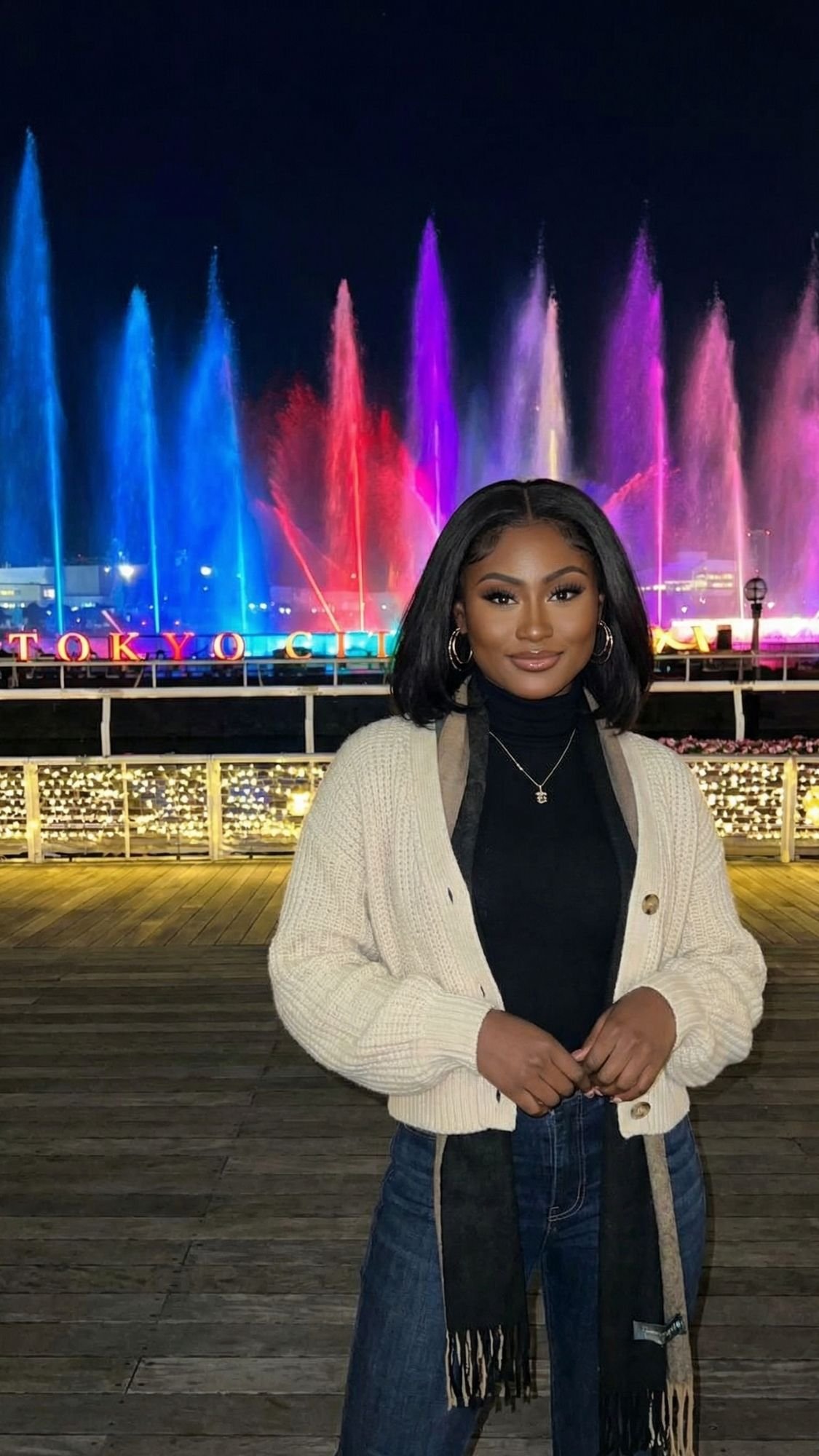 A woman stands on a wooden deck at night, posing in front of a colorful illuminated fountain display. She wears a cream cardigan, black turtleneck, and jeans—enjoying one of the best Tokyo Winter Illuminations with "Tokyo" glowing in the background.