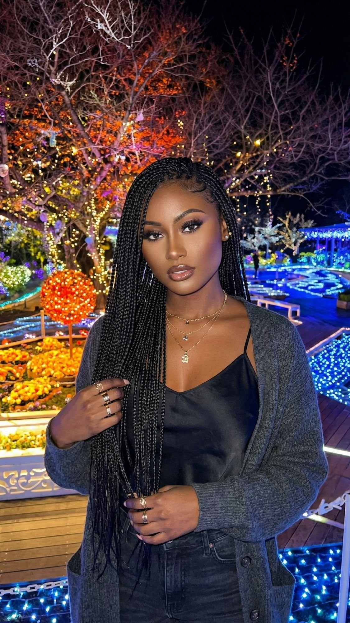 A woman with long braided hair and layered necklaces stands outdoors at night in Tokyo, surrounded by colorful Tokyo Winter Illuminations and glowing trees. In a dark cardigan over a black top, she gazes directly at the camera—perfect for cozy night ideas.