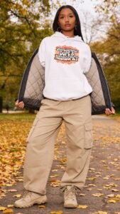 A woman stands on a leaf-covered path in autumn, sporting a cozy fall outfit: a white retro hoodie that reads Unapologetically Dope & Neurospicy, tan cargo pants, beige sneakers, and a black jacket draped open over her shoulders.