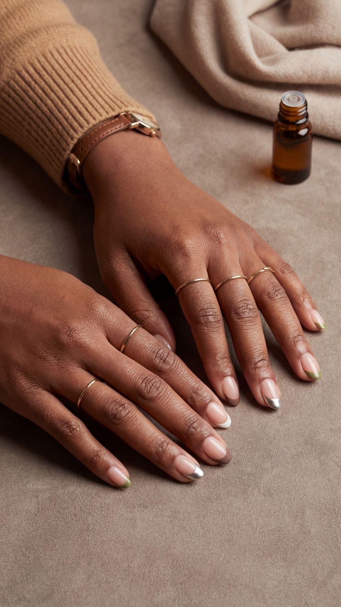 A close-up of two hands with neatly manicured French tip nails and several thin gold rings, resting on a beige surface next to a small amber glass bottle and folded tan fabric, capturing quiet luxury.