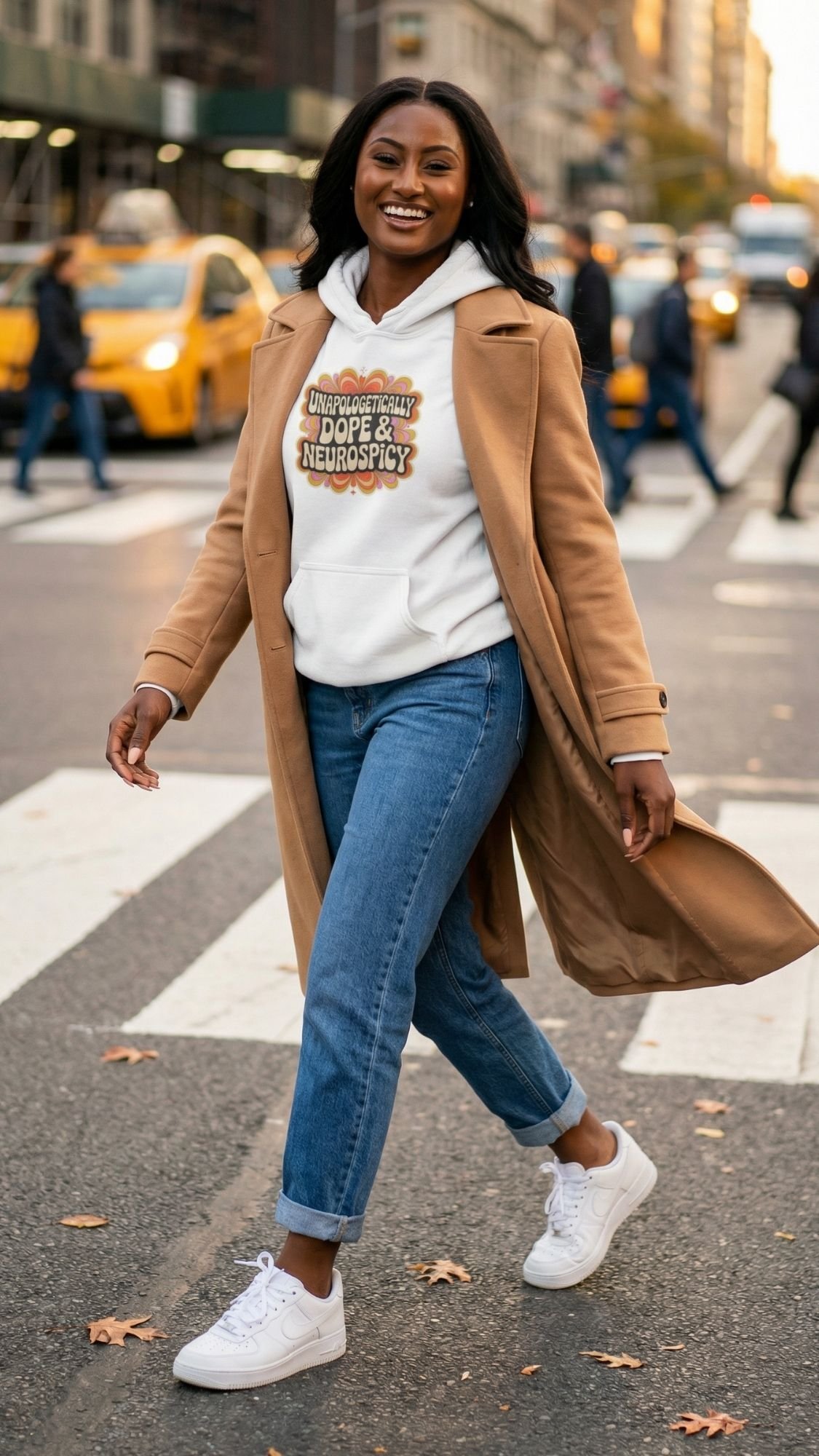 A woman smiles while crossing a city street in a cozy fall outfit: a tan coat over a white retro hoodie with the text Unapologetically Dope & Neurospicy, blue jeans, and white sneakers. Yellow taxis and tall buildings fill the background.