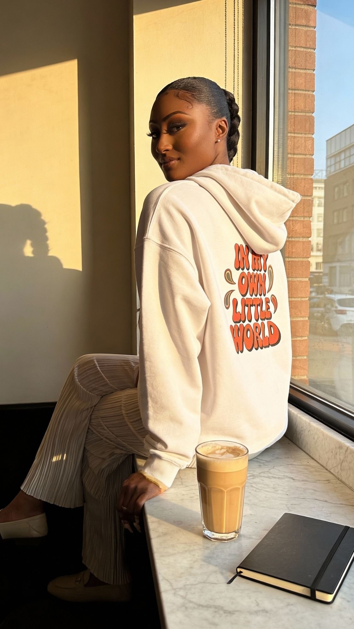 A woman sits on a windowsill, looking over her shoulder. She wears a retro hoodie that reads “IN MY OWN LITTLE WORLD.” A latte and closed notebook rest on the marble ledge, with sunlight streaming in—an effortlessly cozy fall outfit.